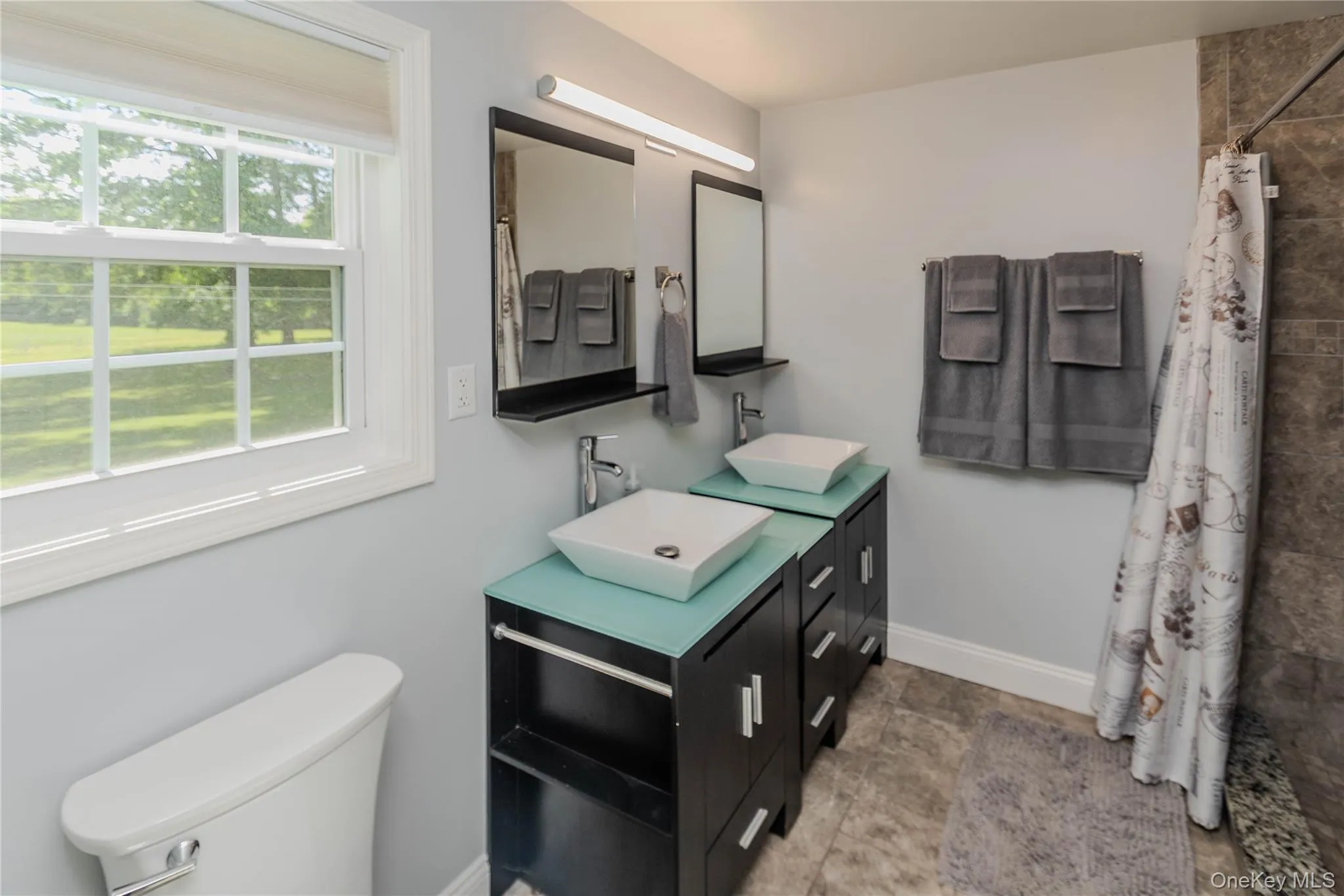 Dual vessel sinks with modern chrome fixtures atop dark wood-finish vanities, complemented by a tiled shower surround and a single-pane window Dual vessel sinks with modern chrome fixtures atop dark wood-finish vanities, complemented by a tiled shower surround and a single-pane window