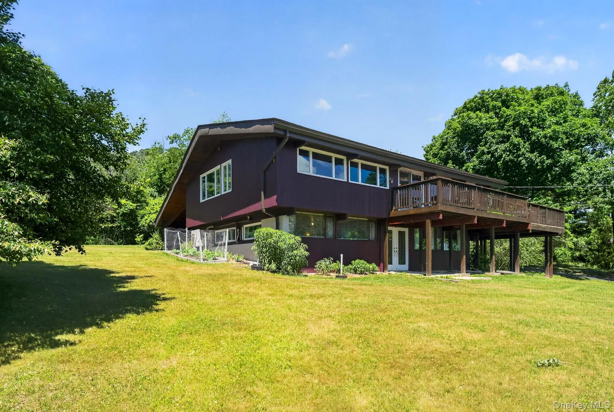 Multi-level exterior with dark siding and white trim, featuring a raised wooden deck, a lower patio, and extensive green lawn space Multi-level exterior with dark siding and white trim, featuring a raised wooden deck, a lower patio, and extensive green lawn space