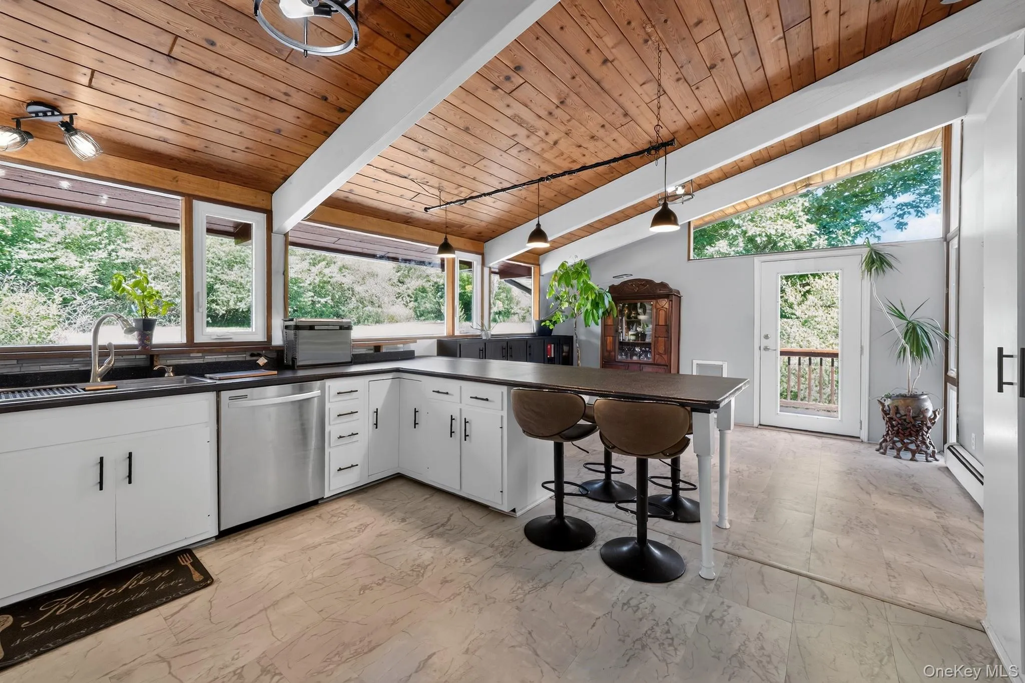 Kitchen featuring a vaulted wood-paneled ceiling, white cabinetry with black hardware, dark countertops, a stainless steel dishwasher, and large windows Kitchen featuring a vaulted wood-paneled ceiling, white cabinetry with black hardware, dark countertops, a stainless steel dishwasher, and large windows