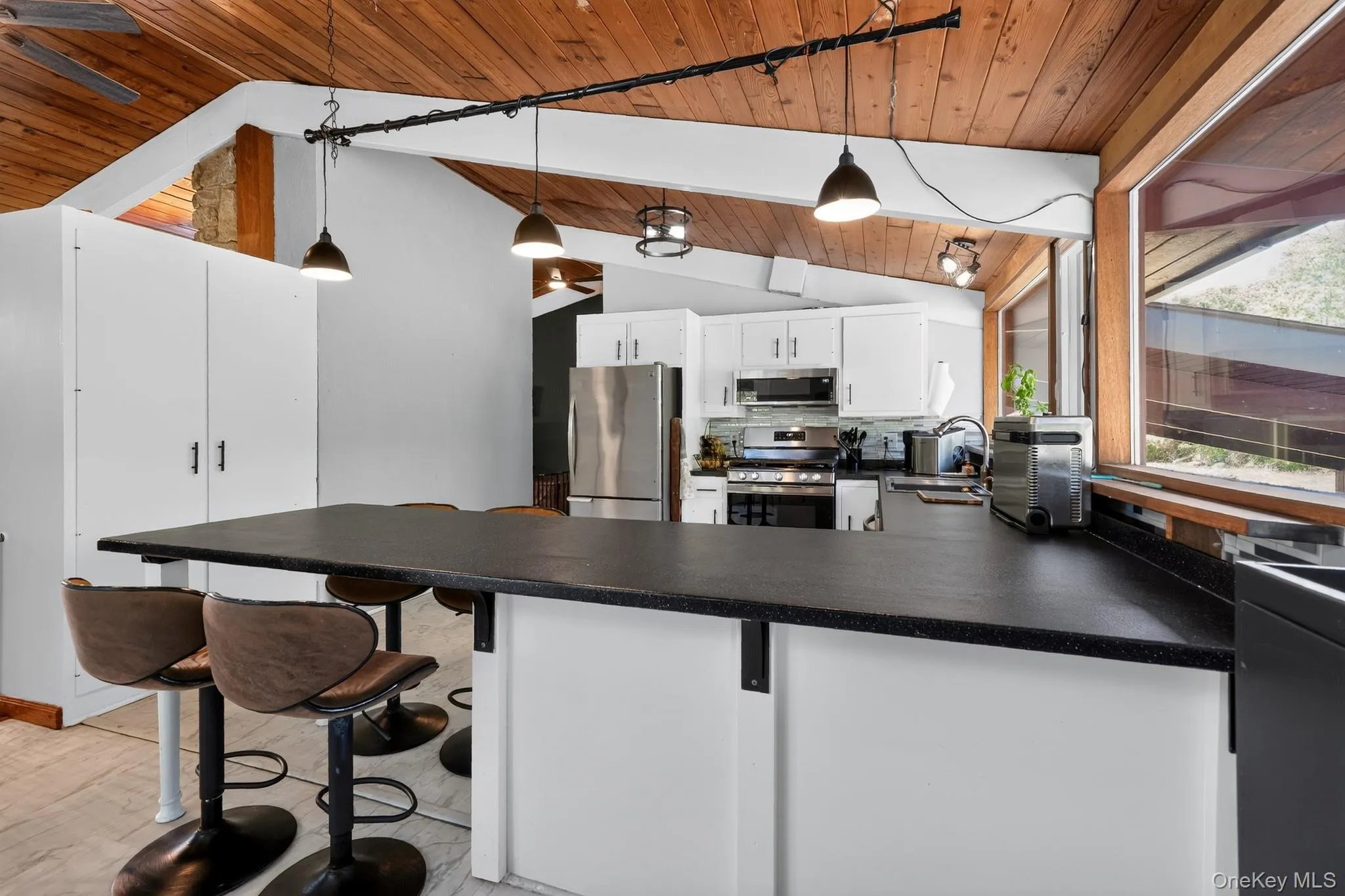Kitchen featuring a long breakfast bar with a solid surface countertop, white cabinetry, stainless steel appliances, and a wood-paneled vaulted ceiling Kitchen featuring a long breakfast bar with a solid surface countertop, white cabinetry, stainless steel appliances, and a wood-paneled vaulted ceiling