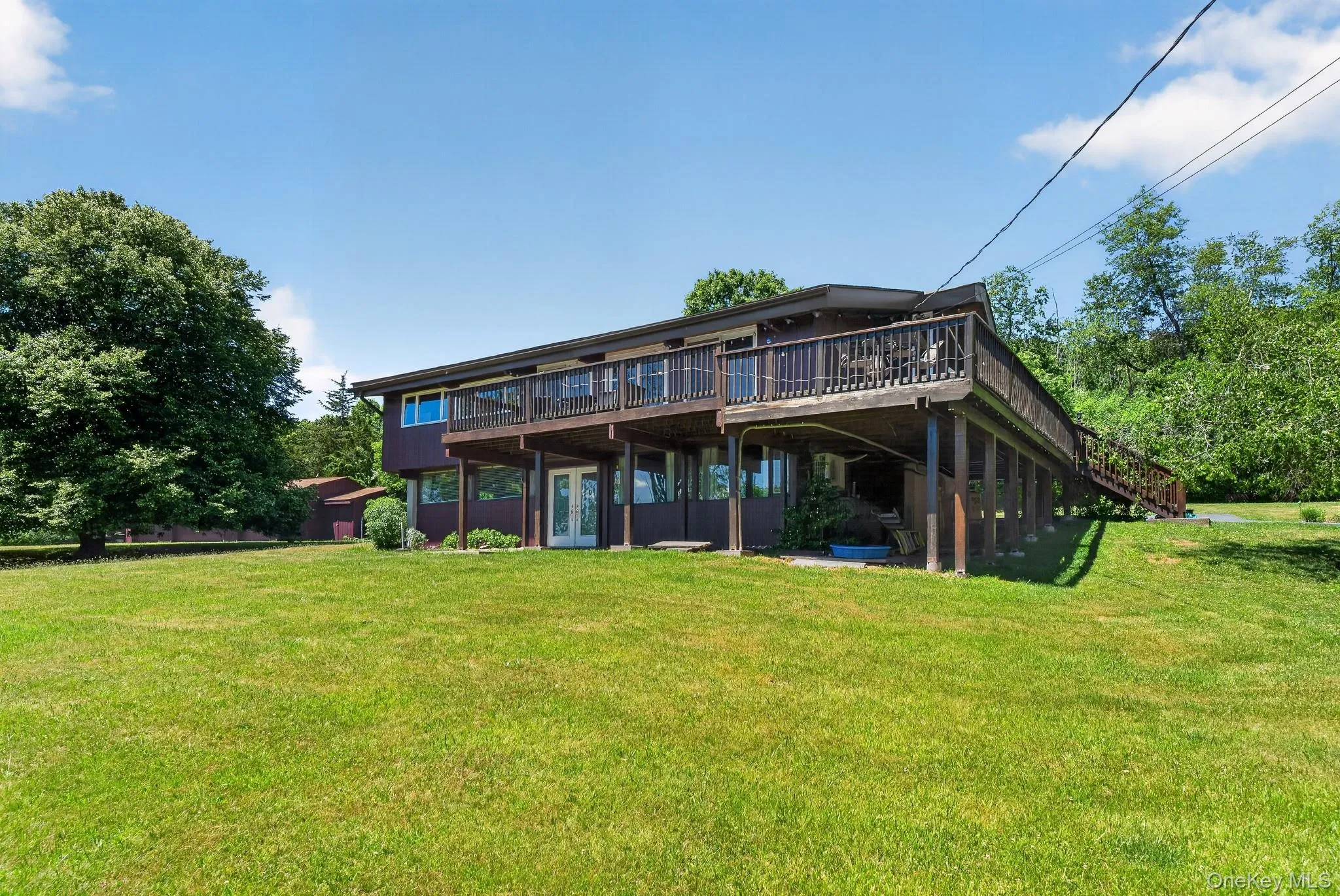 Split-level home featuring an elevated wooden deck spanning the facade, a ground-level patio with double French doors, and an expansive grassy lawn Split-level home featuring an elevated wooden deck spanning the facade, a ground-level patio with double French doors, and an expansive grassy lawn