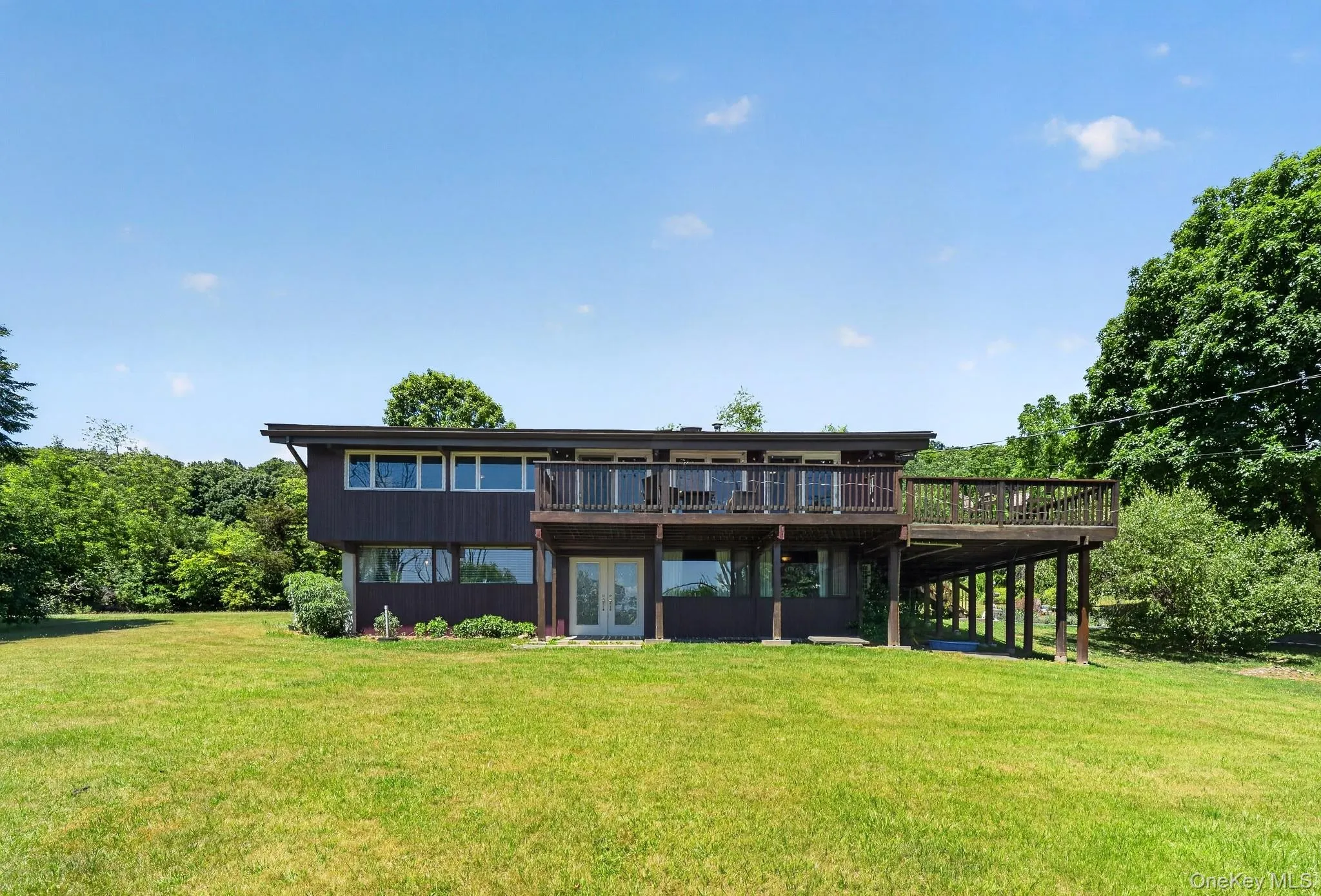 Contemporary two-story residence featuring a dark brown exterior, expansive upper-level deck with wooden railing, and a lower-level patio with French doors Contemporary two-story residence featuring a dark brown exterior, expansive upper-level deck with wooden railing, and a lower-level patio with French doors