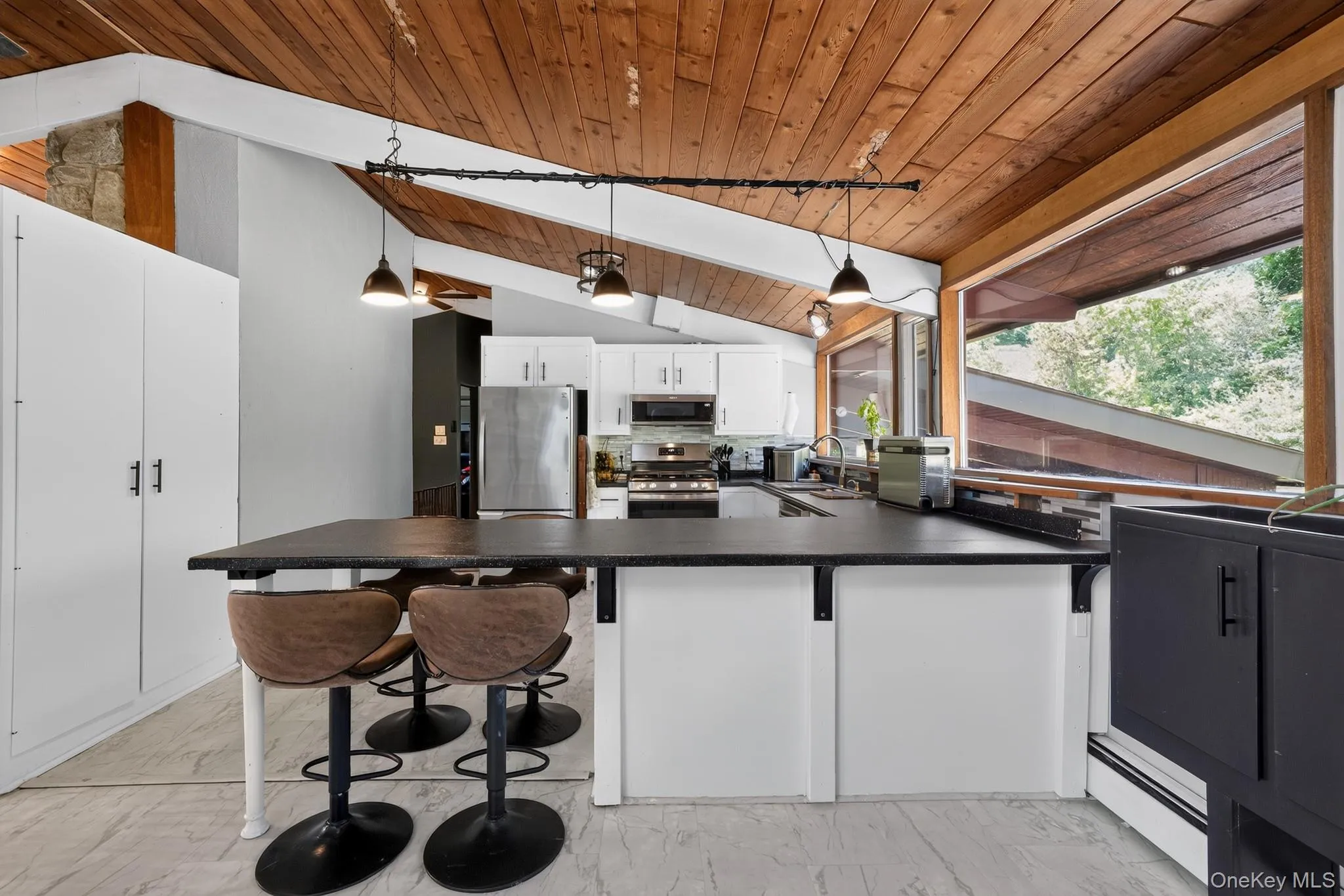 Kitchen featuring an angled wood-panel ceiling, white cabinetry with stainless steel appliances, and a large central island with a dark countertop Kitchen featuring an angled wood-panel ceiling, white cabinetry with stainless steel appliances, and a large central island with a dark countertop