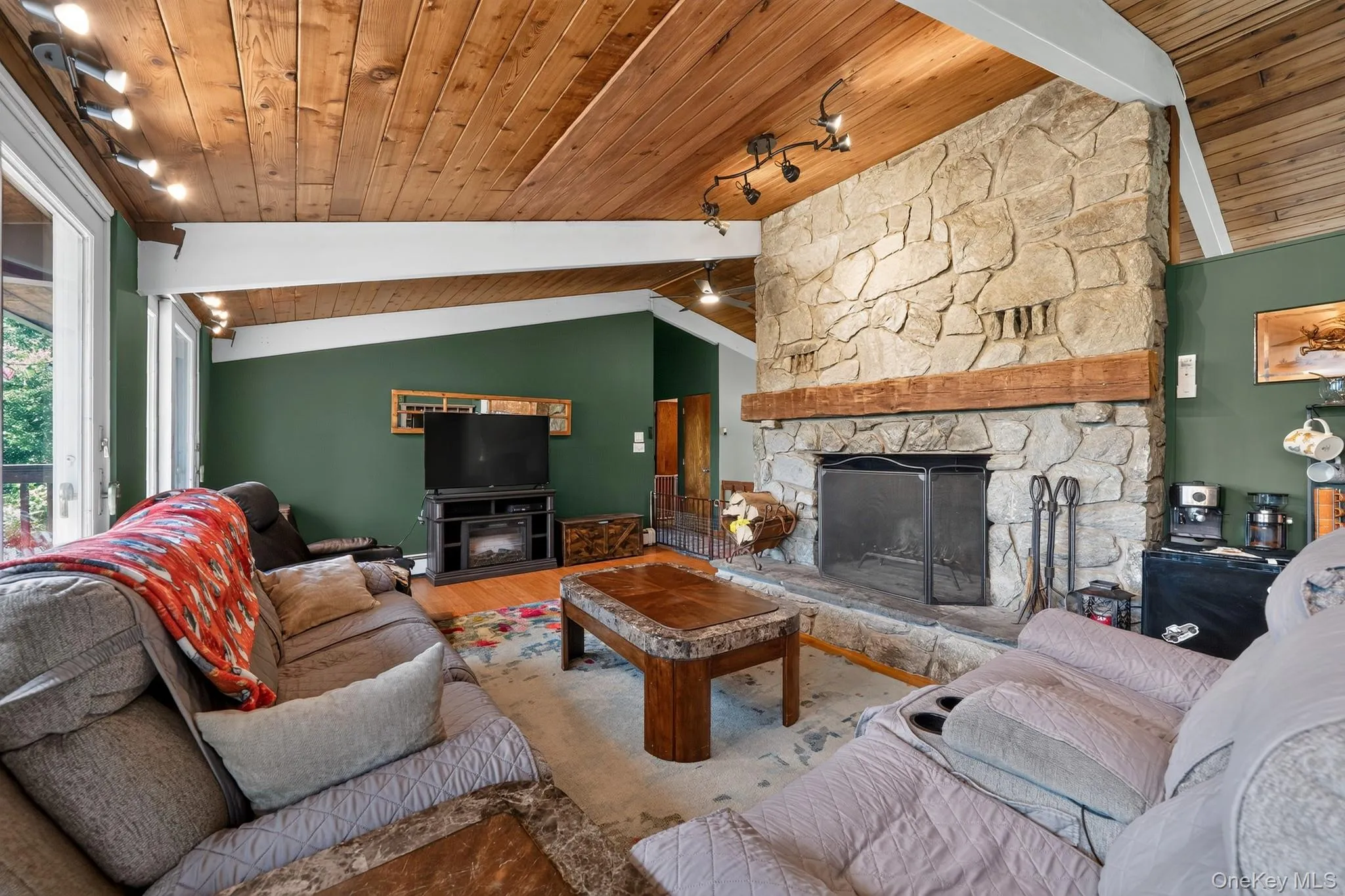 Living area featuring a stone fireplace with a substantial timber mantel, vaulted wood-paneled ceilings, hardwood-style flooring, track lighting, and a glass sliding door Living area featuring a stone fireplace with a substantial timber mantel, vaulted wood-paneled ceilings, hardwood-style flooring, track lighting, and a glass sliding door
