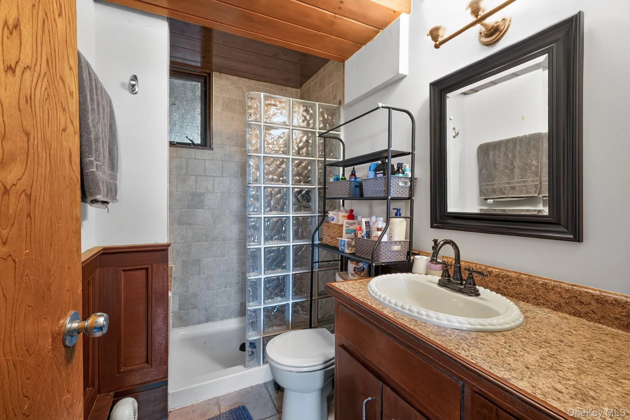 Bathroom featuring a wood-finish ceiling, vanity with a speckled countertop, and a glass block shower enclosure Bathroom featuring a wood-finish ceiling, vanity with a speckled countertop, and a glass block shower enclosure
