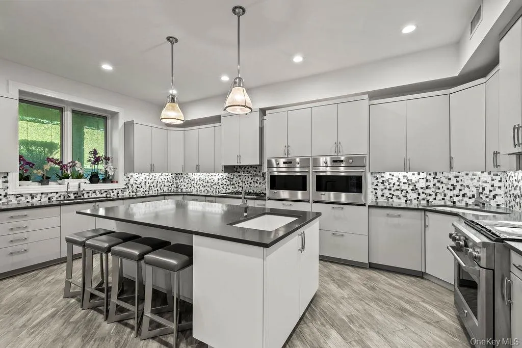 Modern kitchen featuring an expansive center island with an undermount sink, stainless steel appliances, light cabinetry, wood-finish flooring, and a mosaic tile backsplash Modern kitchen featuring an expansive center island with an undermount sink, stainless steel appliances, light cabinetry, wood-finish flooring, and a mosaic tile backsplash