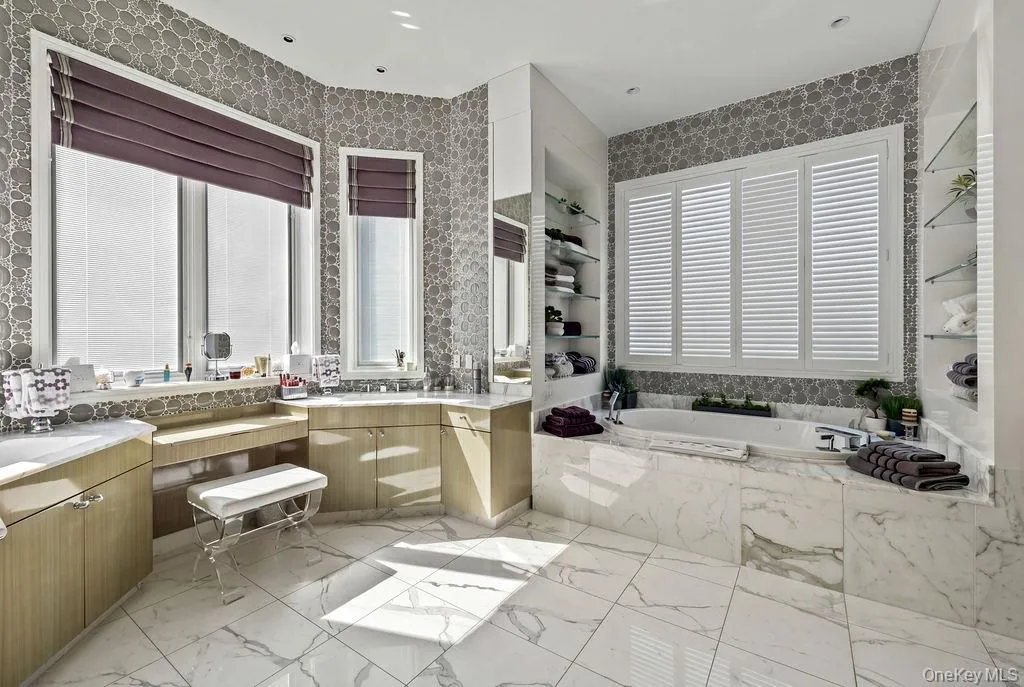 Spacious bathroom featuring large format marble flooring, a built-in soaking tub with marble surround, and an integrated vanity area with dual sinks and light wood-finish cabinetry Spacious bathroom featuring large format marble flooring, a built-in soaking tub with marble surround, and an integrated vanity area with dual sinks and light wood-finish cabinetry