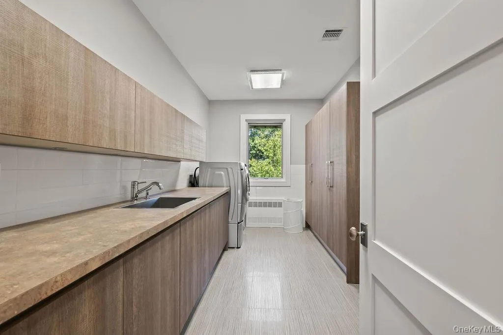 Laundry room featuring extensive wood-finish cabinetry, a deep basin sink with a chrome faucet, light-toned countertop, and a window providing natural illumination Laundry room featuring extensive wood-finish cabinetry, a deep basin sink with a chrome faucet, light-toned countertop, and a window providing natural illumination