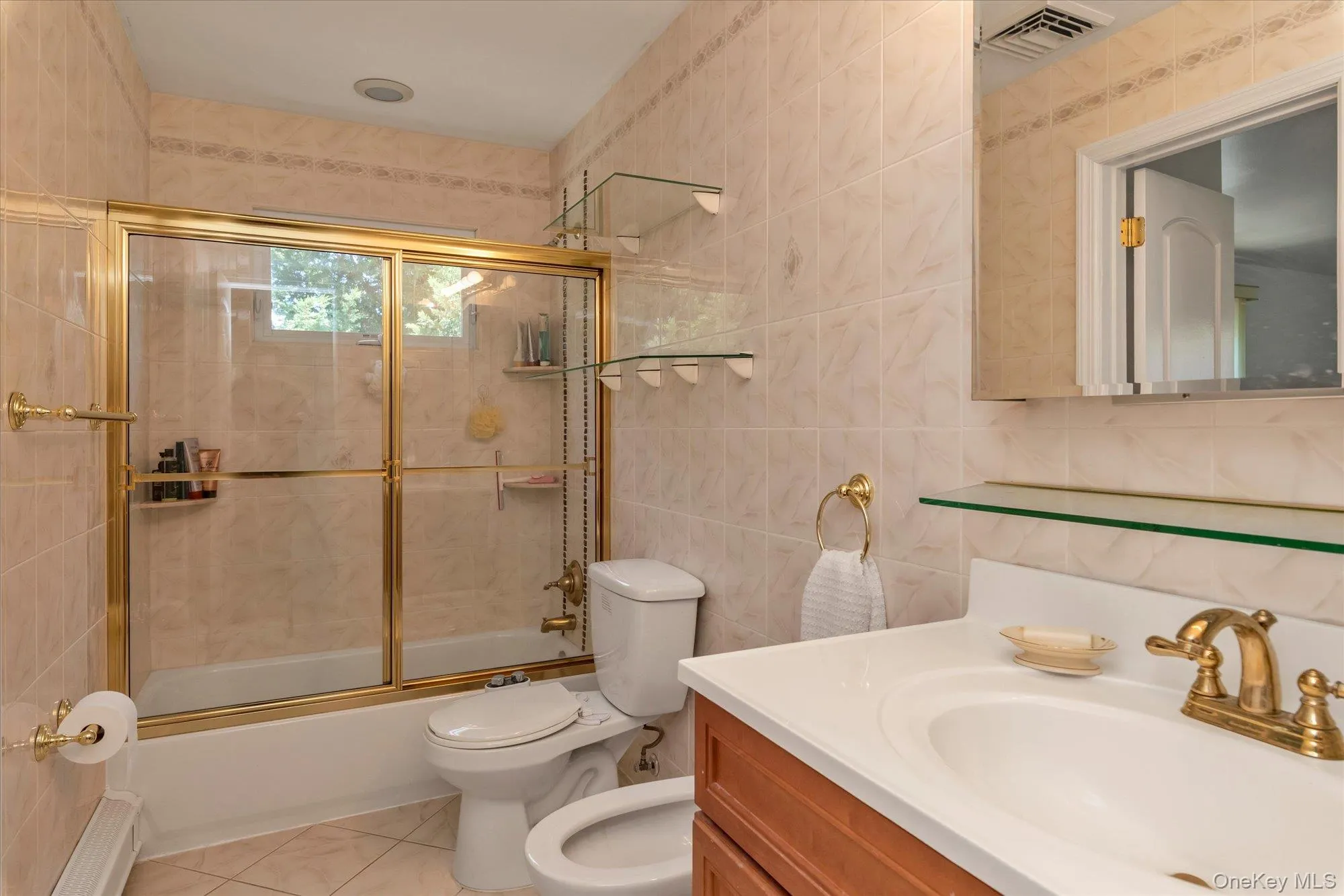 Full bathroom featuring a tub/shower combination with gold-tone framed glass doors, a vanity with a white countertop and integrated sink, gold-tone fixtures, and tiled walls and flooring Full bathroom featuring a tub/shower combination with gold-tone framed glass doors, a vanity with a white countertop and integrated sink, gold-tone fixtures, and tiled walls and flooring