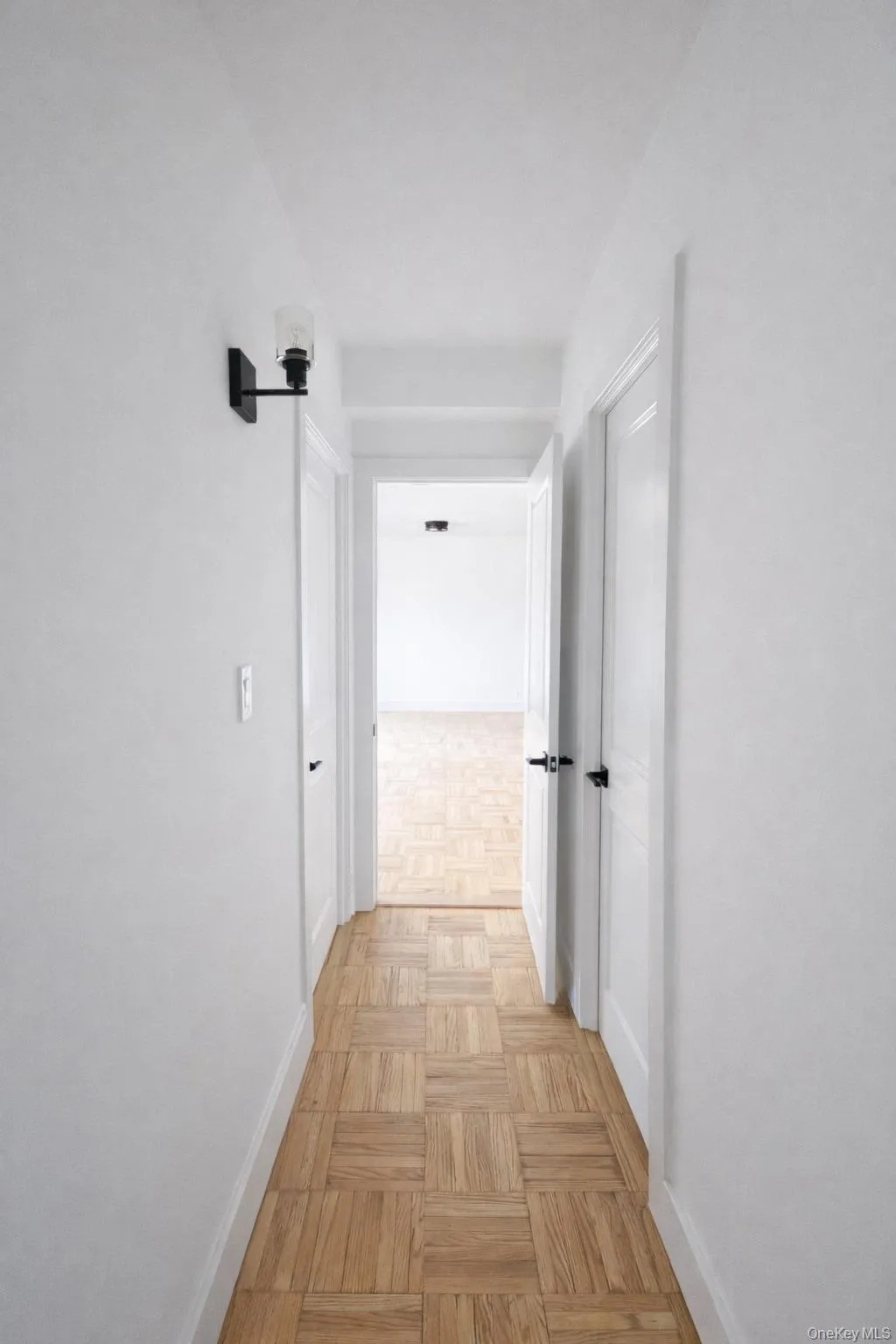 Hallway featuring wood-finish parquet flooring, white walls, and a contemporary wall sconce with a clear glass shade Hallway featuring wood-finish parquet flooring, white walls, and a contemporary wall sconce with a clear glass shade