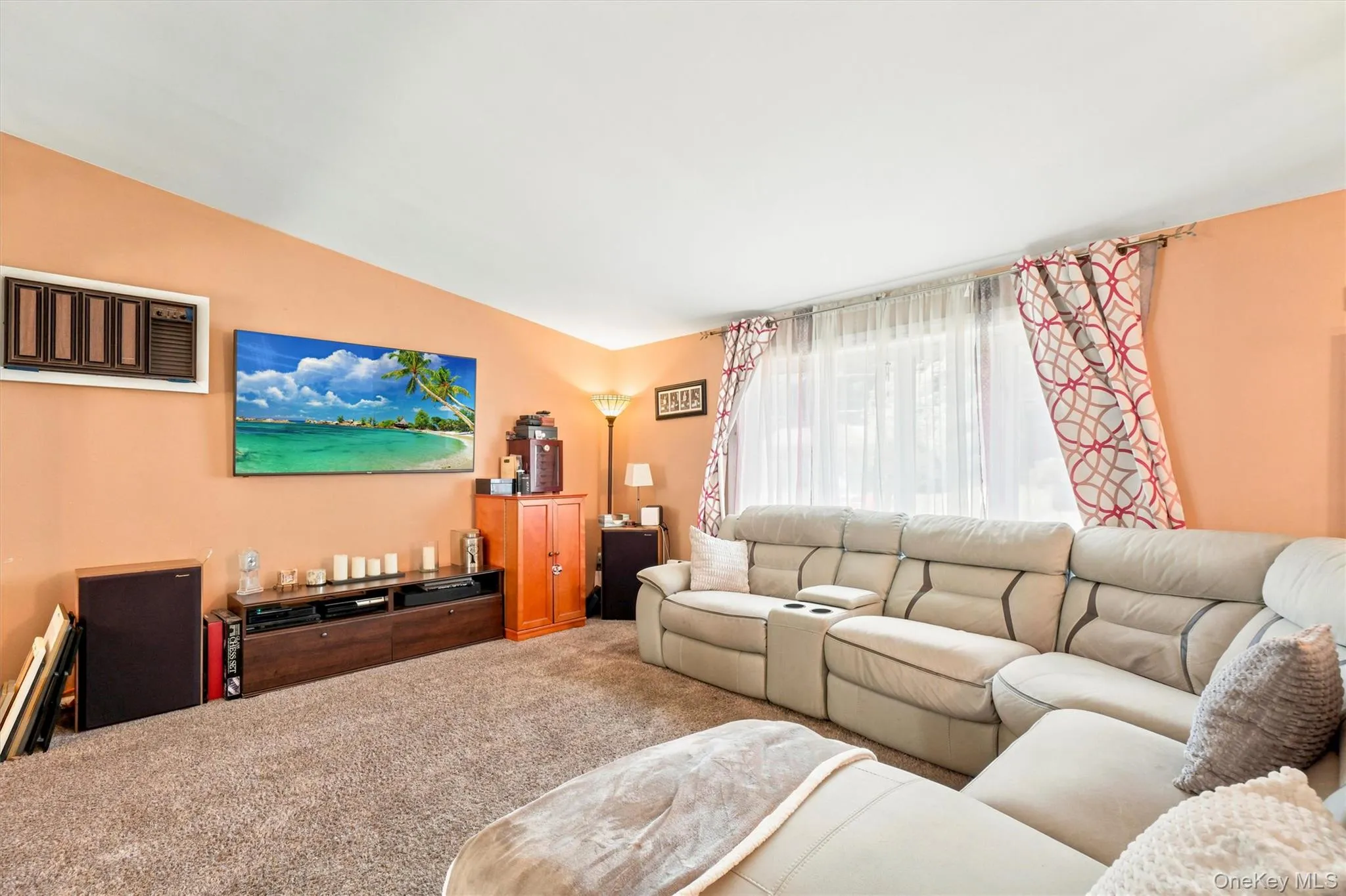 Living area featuring neutral-toned carpeting, a window with sheer and patterned drapes, a wall-mounted air conditioning unit, and a built-in media console Living area featuring neutral-toned carpeting, a window with sheer and patterned drapes, a wall-mounted air conditioning unit, and a built-in media console