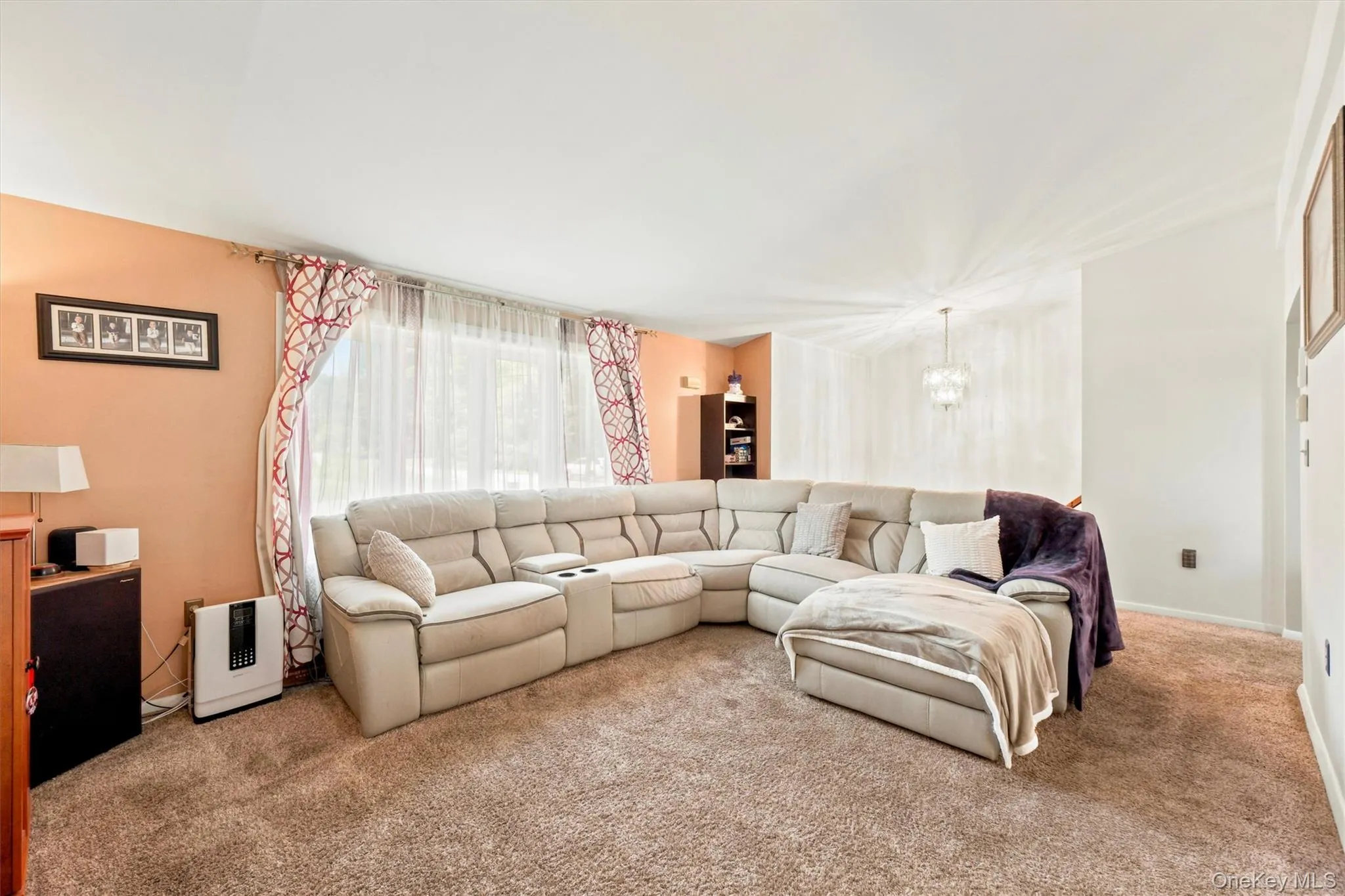 Carpeted living area featuring a large window with sheer and patterned curtains, light-colored walls, and a built-in shelving unit Carpeted living area featuring a large window with sheer and patterned curtains, light-colored walls, and a built-in shelving unit
