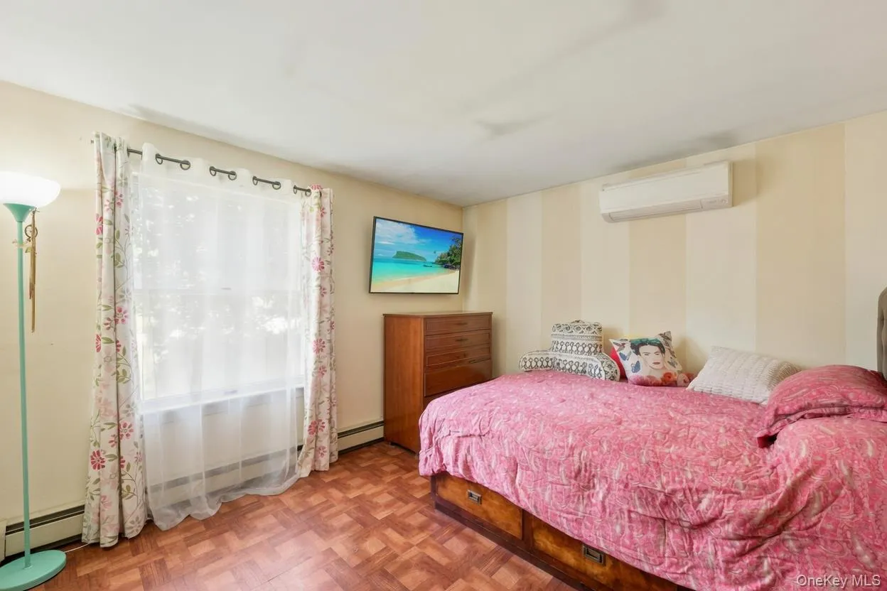 Room with wood-finish parquet flooring, a wall-mounted AC unit, and a single window with curtain rods Room with wood-finish parquet flooring, a wall-mounted AC unit, and a single window with curtain rods