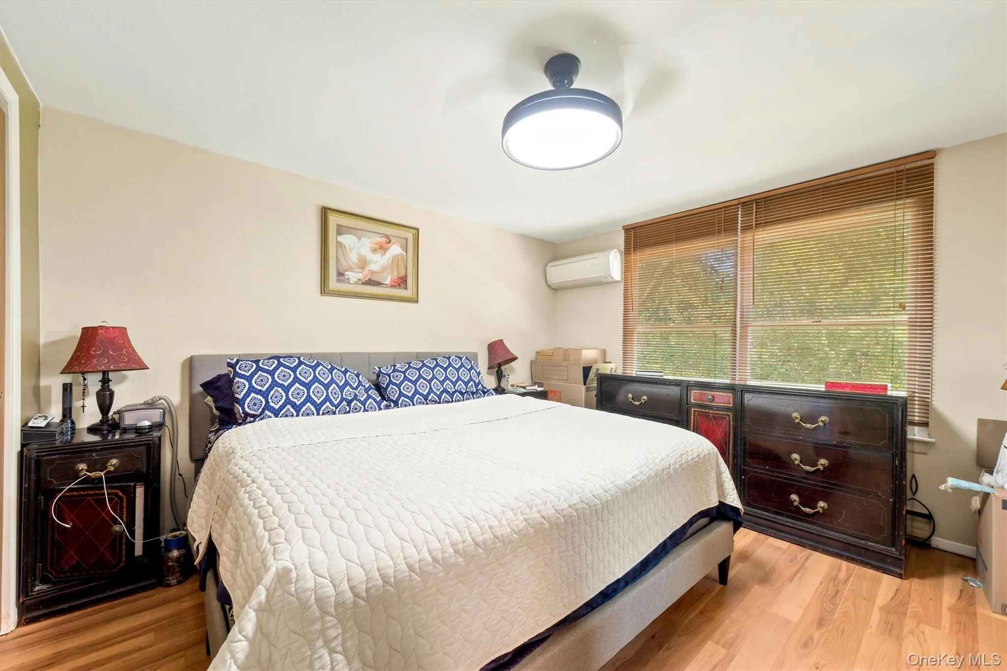 Bedroom featuring wood-finish flooring, a double window with horizontal blinds, and a wall-mounted air conditioning unit Bedroom featuring wood-finish flooring, a double window with horizontal blinds, and a wall-mounted air conditioning unit
