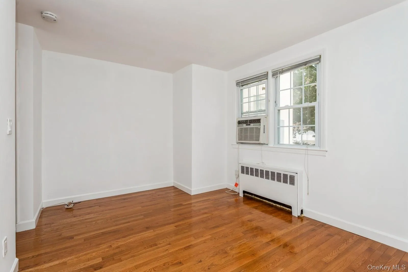 Unfurnished room featuring radiator, a smoke detector, wood finished floors, and cooling unit Unfurnished room featuring radiator, a smoke detector, wood finished floors, and cooling unit