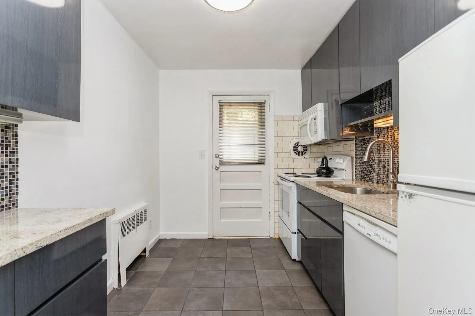 Kitchen featuring white appliances, tasteful backsplash, radiator, dark tile patterned flooring, and light stone counters Kitchen featuring white appliances, tasteful backsplash, radiator, dark tile patterned flooring, and light stone counters