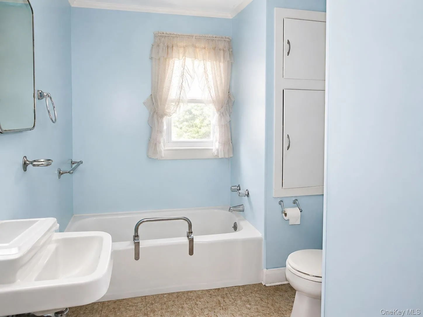 Bathroom featuring a window, white bathtub, wall-mounted sink, built-in cabinetry, and ceramic toilet Bathroom featuring a window, white bathtub, wall-mounted sink, built-in cabinetry, and ceramic toilet