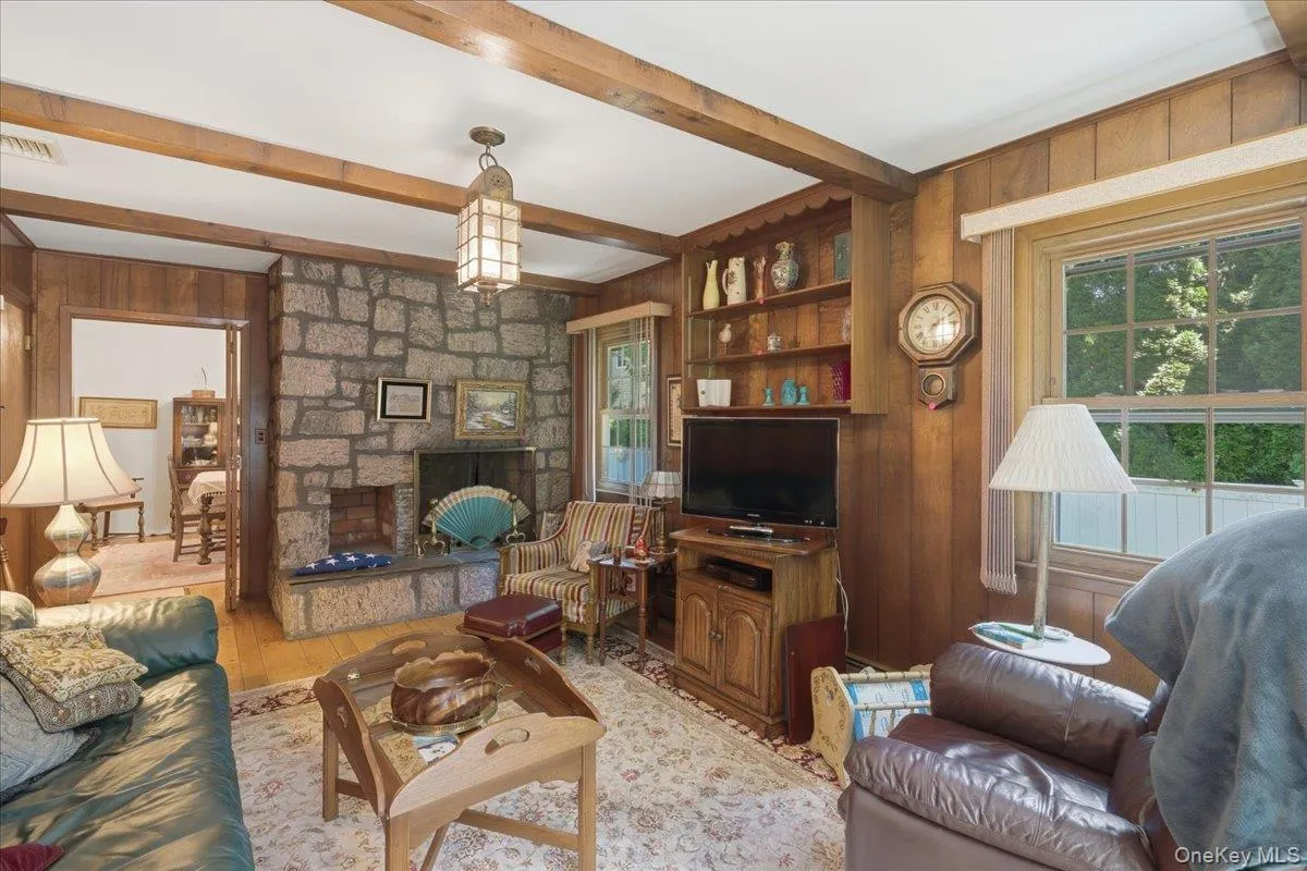 Living space featuring a stone fireplace with a raised hearth, wood-finish wall paneling, exposed ceiling beams, built-in shelving, and wood-finish flooring Living space featuring a stone fireplace with a raised hearth, wood-finish wall paneling, exposed ceiling beams, built-in shelving, and wood-finish flooring