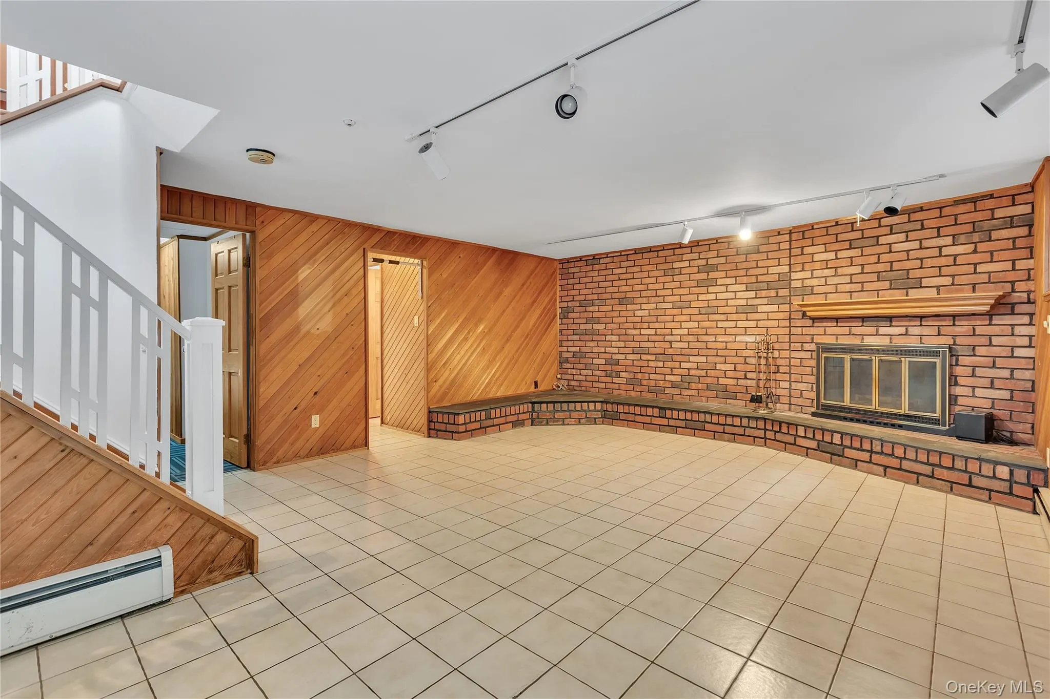 Spacious open-concept room featuring a brick-accented wall with a fireplace, a wood-paneled accent wall, and tiled flooring Spacious open-concept room featuring a brick-accented wall with a fireplace, a wood-paneled accent wall, and tiled flooring