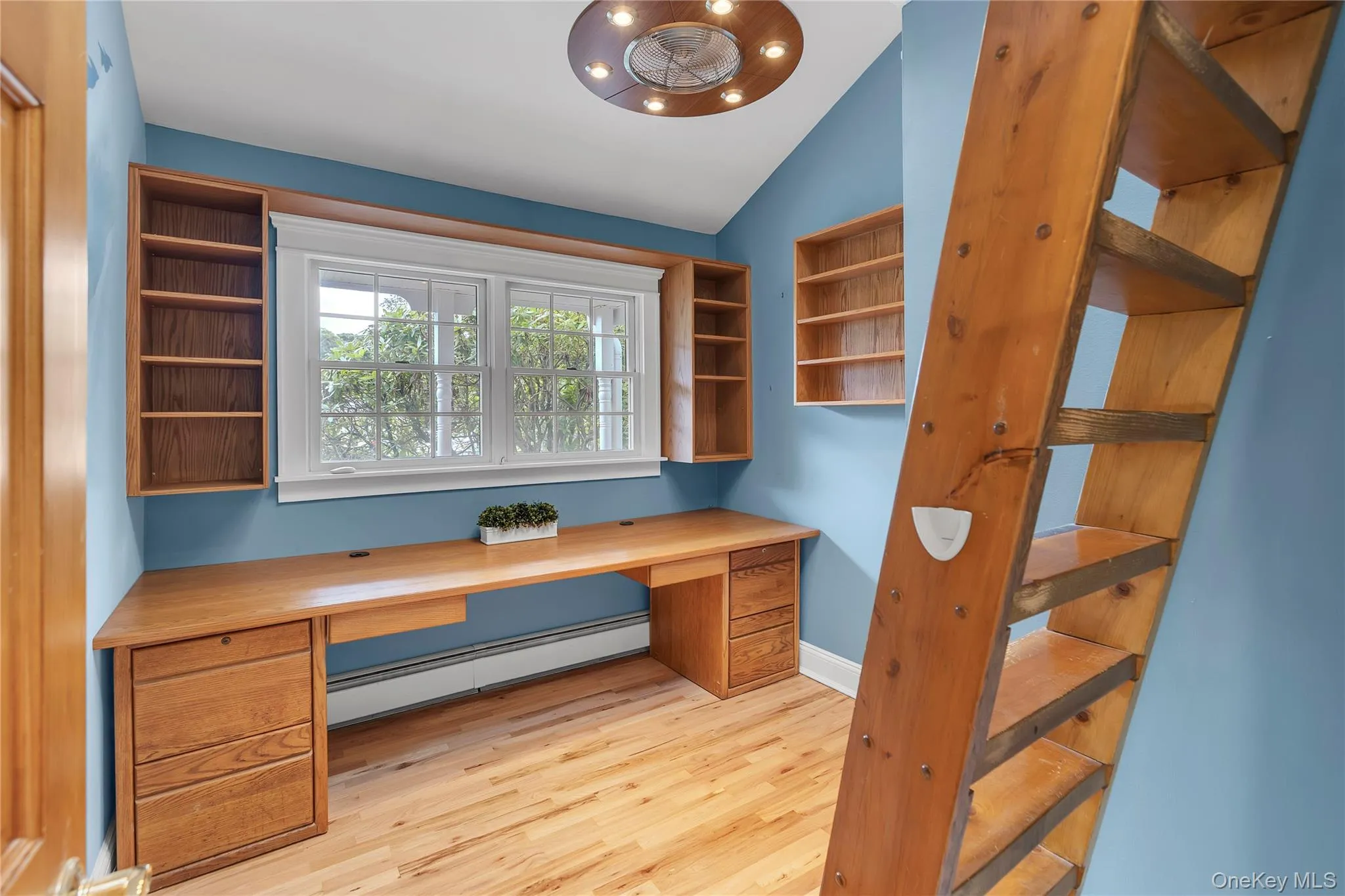 Dedicated workspace featuring a built-in wood desk with integrated drawers, wood-finish flooring, and a large window providing natural light. Stairs with loft with bed Dedicated workspace featuring a built-in wood desk with integrated drawers, wood-finish flooring, and a large window providing natural light. Stairs with loft with bed