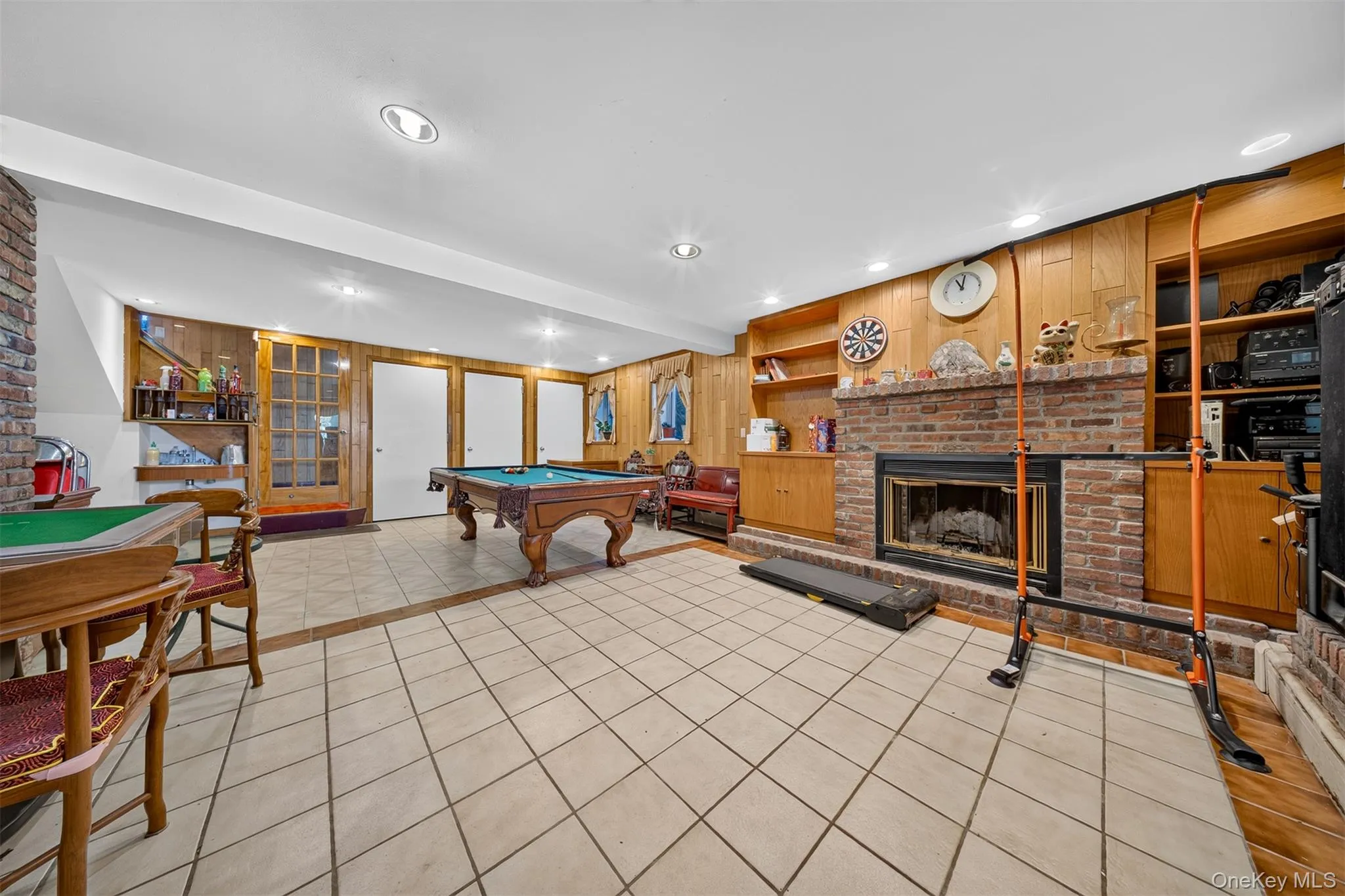 Spacious entertainment area featuring tile flooring, a brick fireplace with a hearth, wood paneling, recessed lighting, and built-in shelving Spacious entertainment area featuring tile flooring, a brick fireplace with a hearth, wood paneling, recessed lighting, and built-in shelving