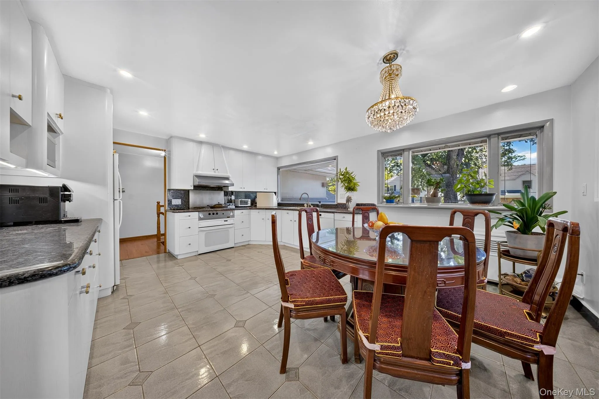 Bright kitchen and dining area featuring tile flooring, white cabinetry, dark stone countertops, and a crystal chandelier Bright kitchen and dining area featuring tile flooring, white cabinetry, dark stone countertops, and a crystal chandelier