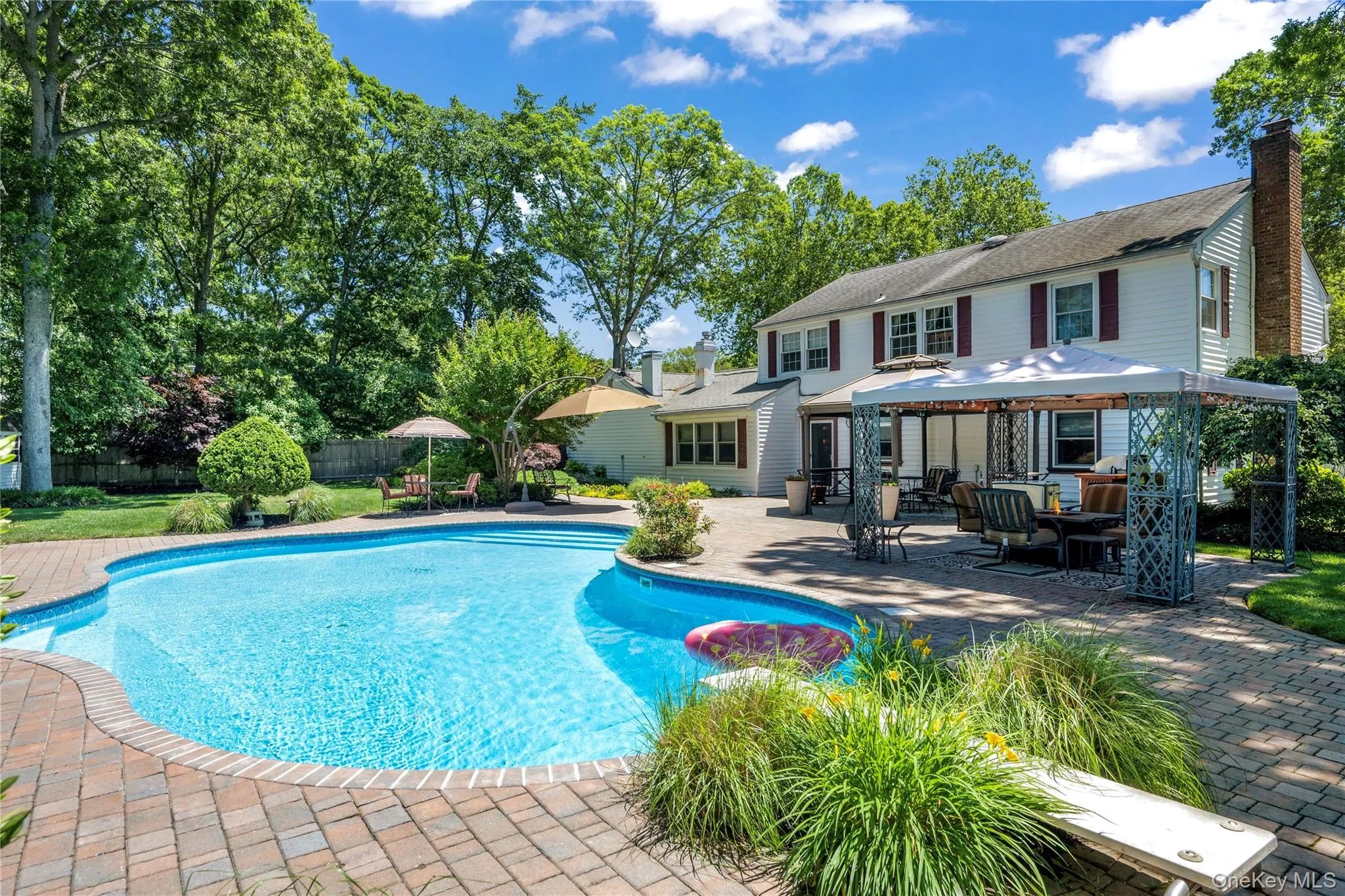 Rear exterior featuring a curved inground swimming pool with brick paver surround, an expansive paver patio with a gazebo structure, and mature trees providing a lush backdrop Rear exterior featuring a curved inground swimming pool with brick paver surround, an expansive paver patio with a gazebo structure, and mature trees providing a lush backdrop