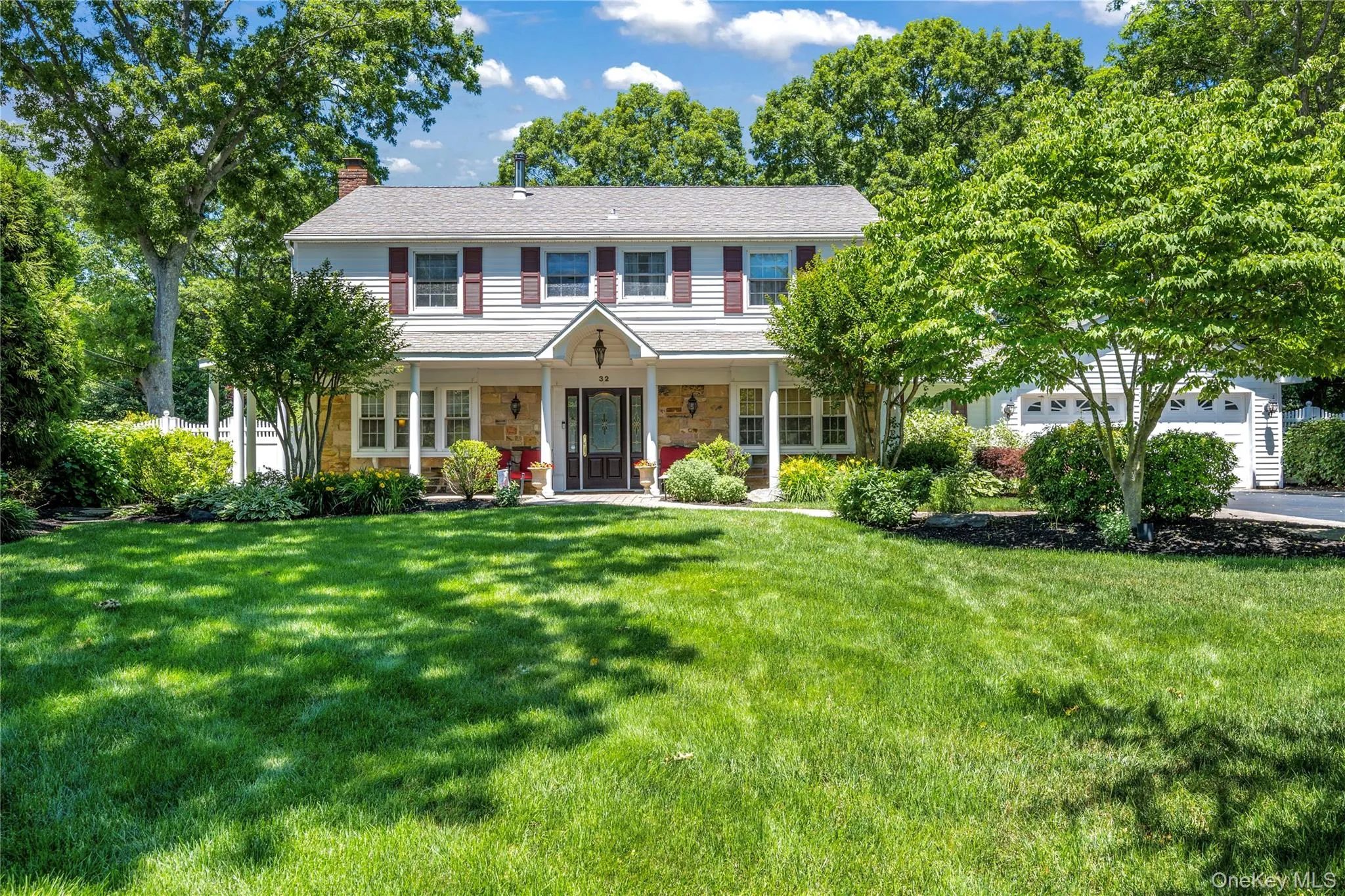 Two-story traditional Colonial featuring a full-width covered porch with white columns, a dark wood entry door with glass inserts, and contrasting burgundy shutters Two-story traditional Colonial featuring a full-width covered porch with white columns, a dark wood entry door with glass inserts, and contrasting burgundy shutters