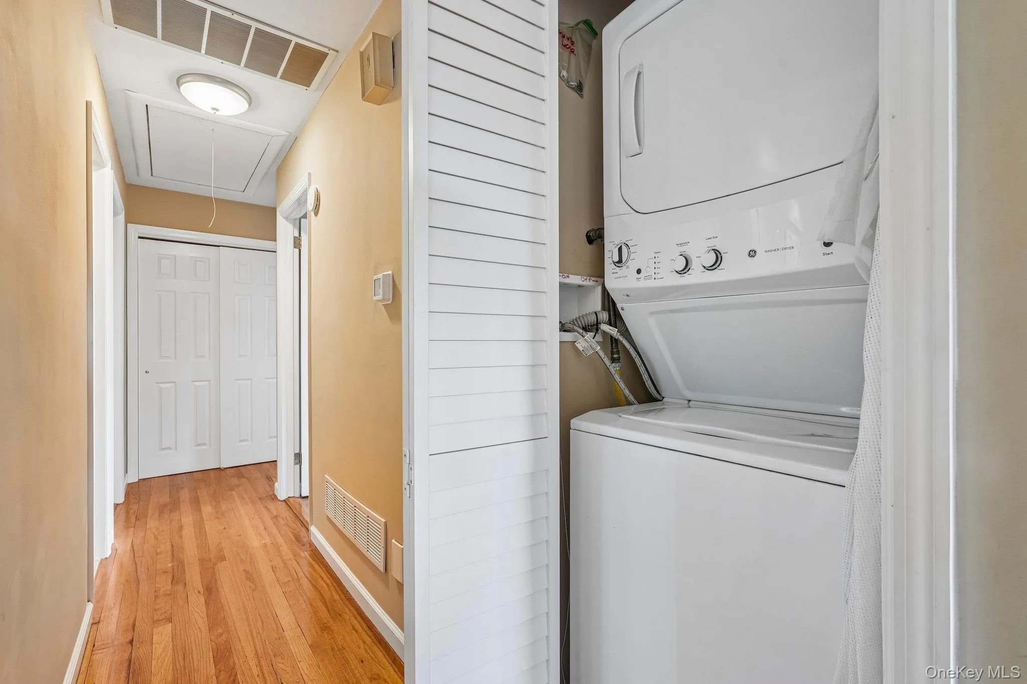 Hallway laundry closet with a stacked washer and dryer, louvered bi-fold doors, and wood-finish flooring Hallway laundry closet with a stacked washer and dryer, louvered bi-fold doors, and wood-finish flooring