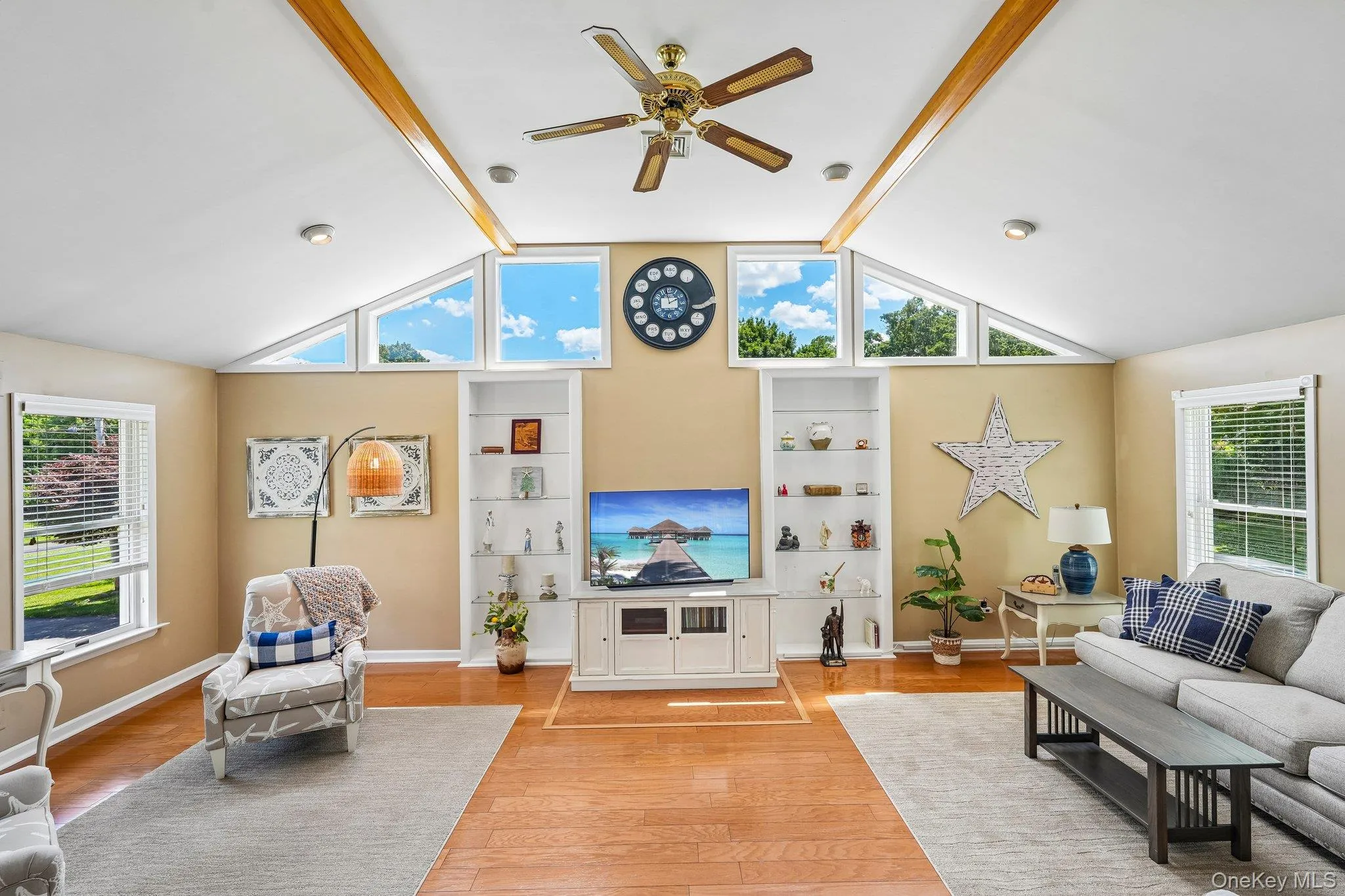 Vaulted ceiling living area featuring wood-finish flooring, exposed ceiling beams, a ceiling fan, and multiple windows including high transom panes Vaulted ceiling living area featuring wood-finish flooring, exposed ceiling beams, a ceiling fan, and multiple windows including high transom panes