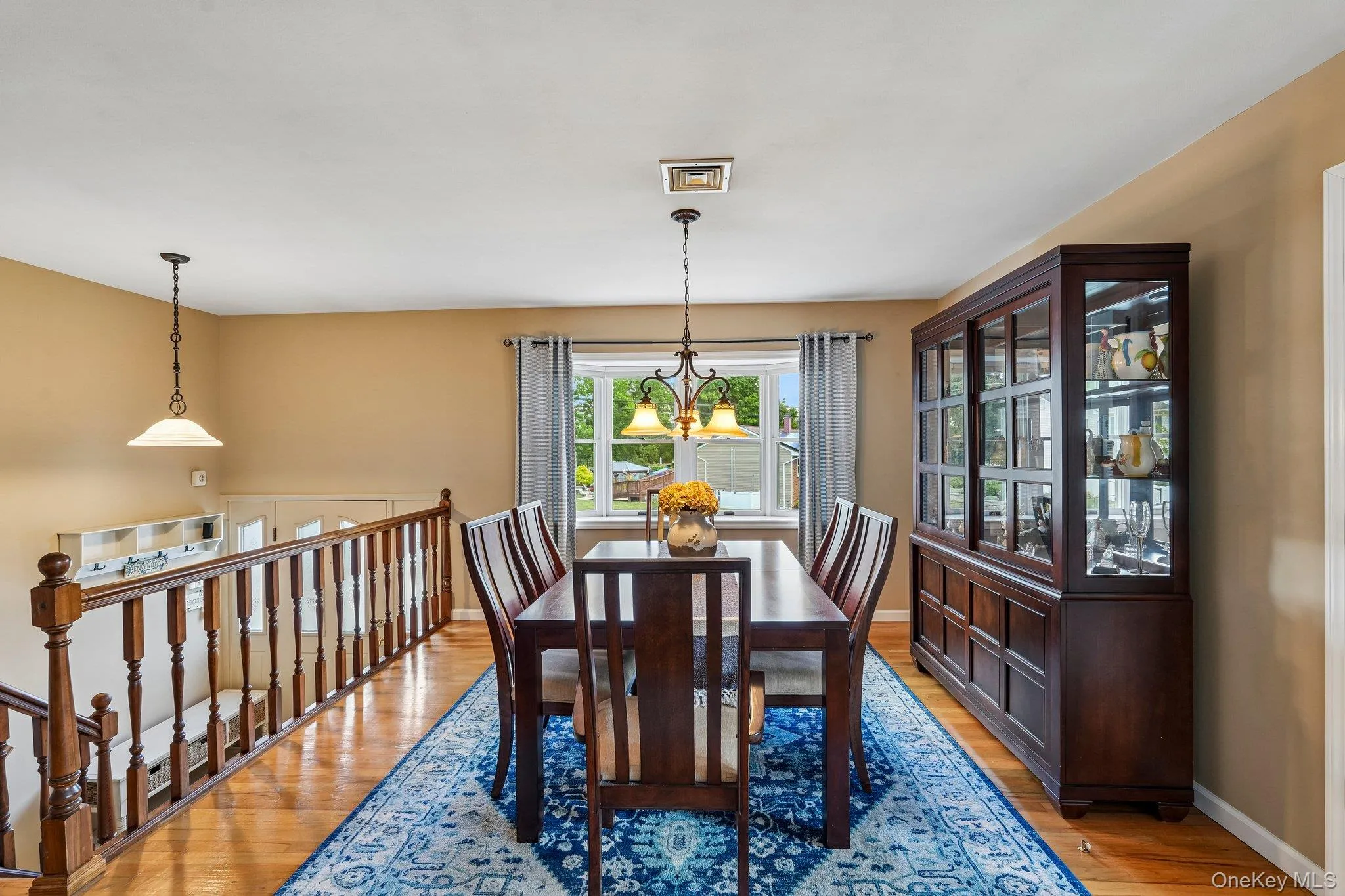 Bright dining area featuring hardwood flooring, a bay window with natural light, a wood spindle railing, and a decorative chandelier Bright dining area featuring hardwood flooring, a bay window with natural light, a wood spindle railing, and a decorative chandelier