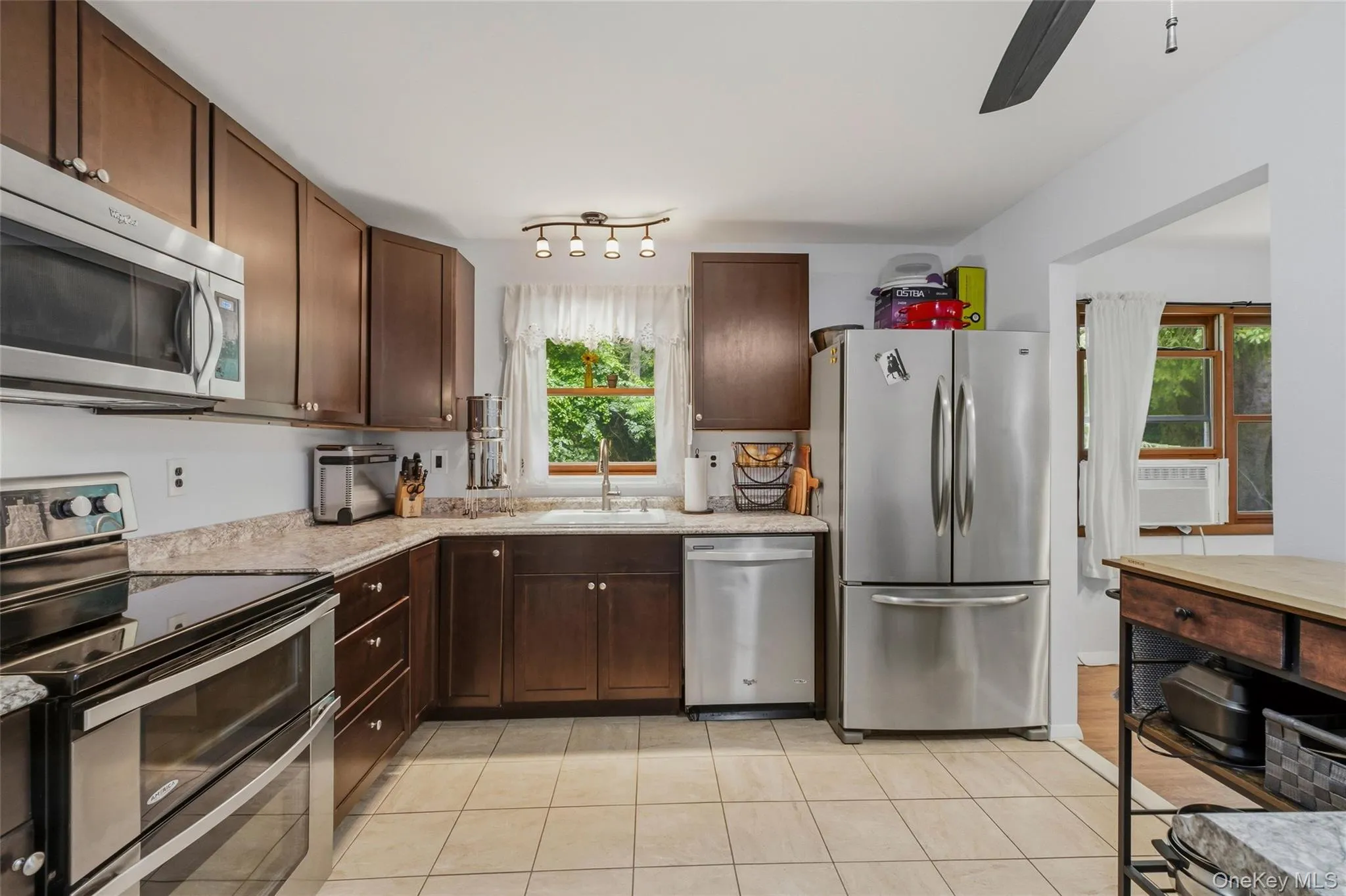 Modern kitchen featuring rich wood cabinetry, light-toned countertops, and a full suite of stainless steel appliances and window Modern kitchen featuring rich wood cabinetry, light-toned countertops, and a full suite of stainless steel appliances and window