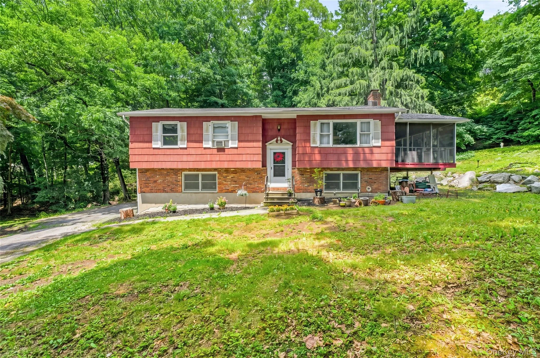 Raised ranch house featuring a red siding and brick facade, a screened-in porch, a paved driveway, and mature landscaping Raised ranch house featuring a red siding and brick facade, a screened-in porch, a paved driveway, and mature landscaping