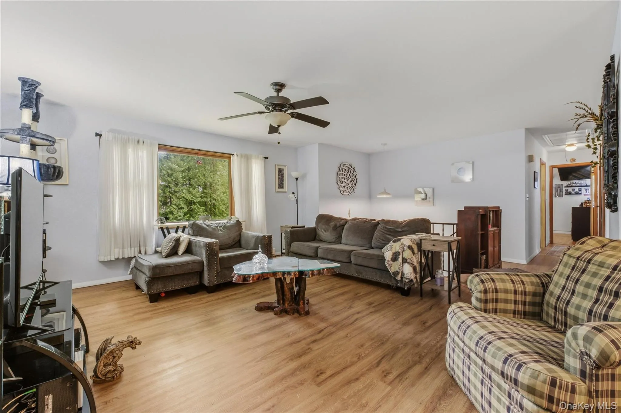 Spacious living area featuring wood-finish flooring, light-toned walls, and a ceiling fan with a large window. Spacious living area featuring wood-finish flooring, light-toned walls, and a ceiling fan with a large window.