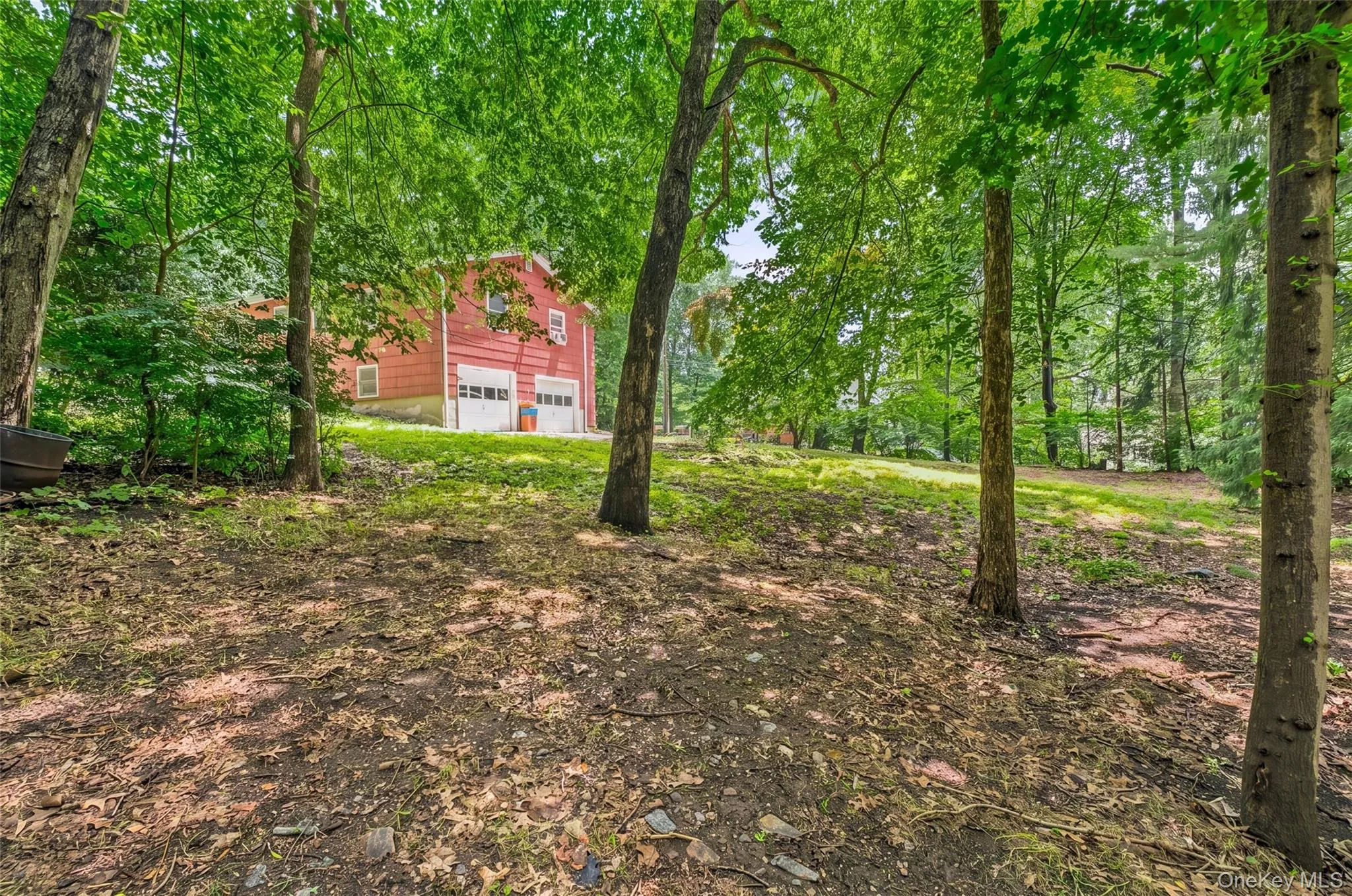 Red-side raised ranch structure featuring two white garage doors and upper-level windows, situated on a sloped, tree-lined lot Red-side raised ranch structure featuring two white garage doors and upper-level windows, situated on a sloped, tree-lined lot