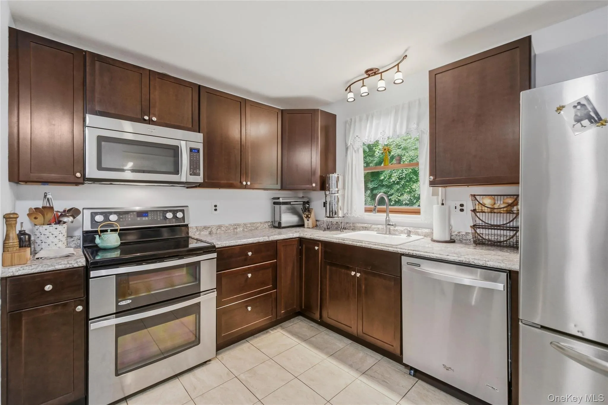 Modern kitchen featuring rich wood cabinetry, light-toned countertops, and a full suite of stainless steel appliances and window Modern kitchen featuring rich wood cabinetry, light-toned countertops, and a full suite of stainless steel appliances and window