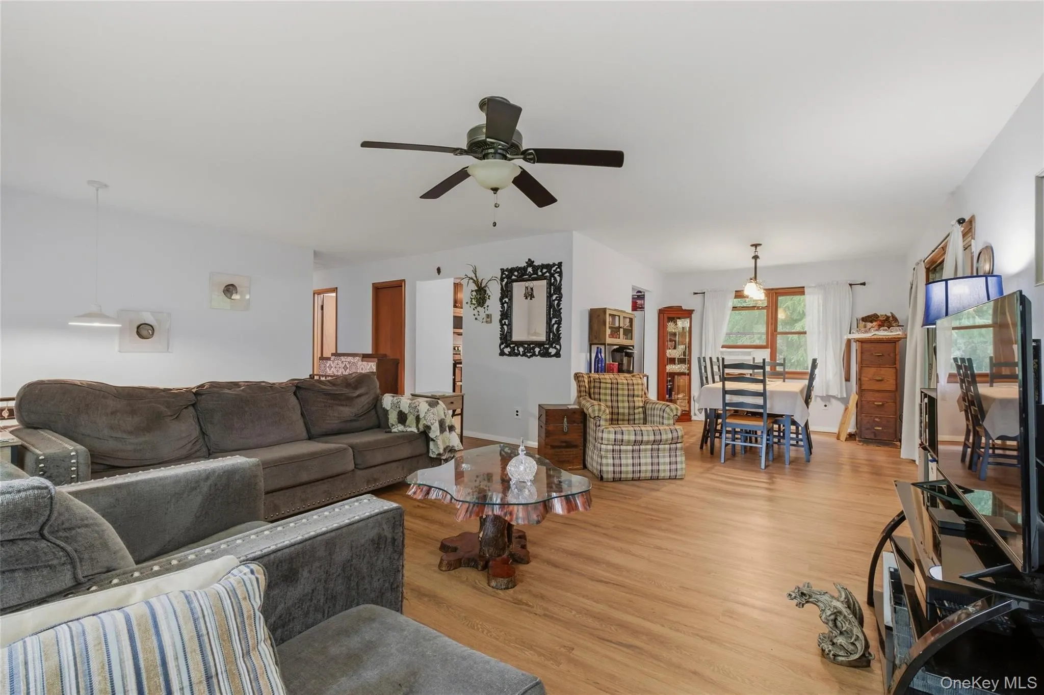 Spacious living area featuring wood-finish flooring, a ceiling fan, and white walls leading to a dining area Spacious living area featuring wood-finish flooring, a ceiling fan, and white walls leading to a dining area