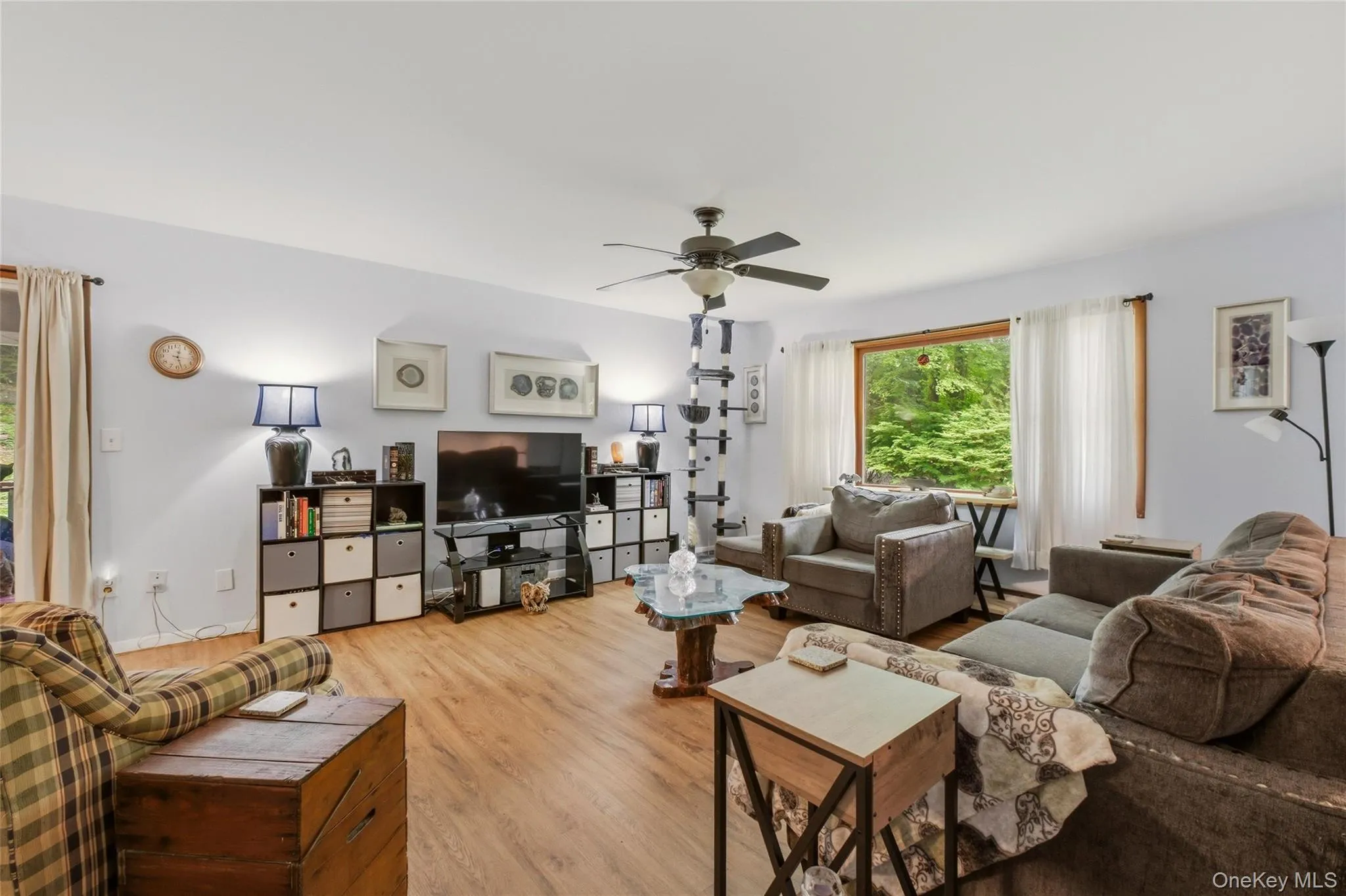 Spacious living area featuring wood-finish flooring, light-toned walls, and a ceiling fan with a large window. Spacious living area featuring wood-finish flooring, light-toned walls, and a ceiling fan with a large window.