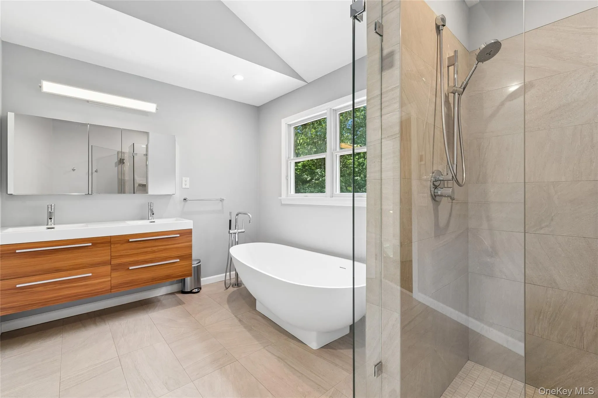 Contemporary bathroom featuring a frameless glass shower enclosure, a freestanding soaking tub with a floor-mounted faucet, a floating double vanity with integrated sinks, and neutral-toned floor and shower wall tiling Contemporary bathroom featuring a frameless glass shower enclosure, a freestanding soaking tub with a floor-mounted faucet, a floating double vanity with integrated sinks, and neutral-toned floor and shower wall tiling