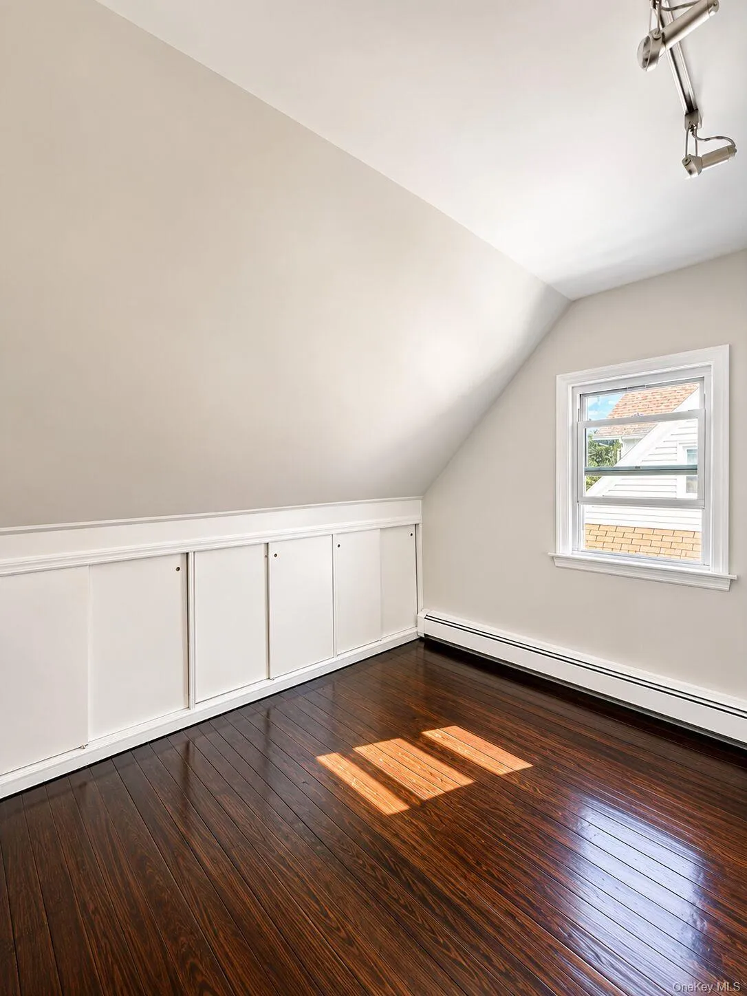 Second bedroom featuring wood-finish flooring, built-in lower cabinetry with paneling, a single window with white trim, a sloped ceiling, and baseboard heating Second bedroom featuring wood-finish flooring, built-in lower cabinetry with paneling, a single window with white trim, a sloped ceiling, and baseboard heating
