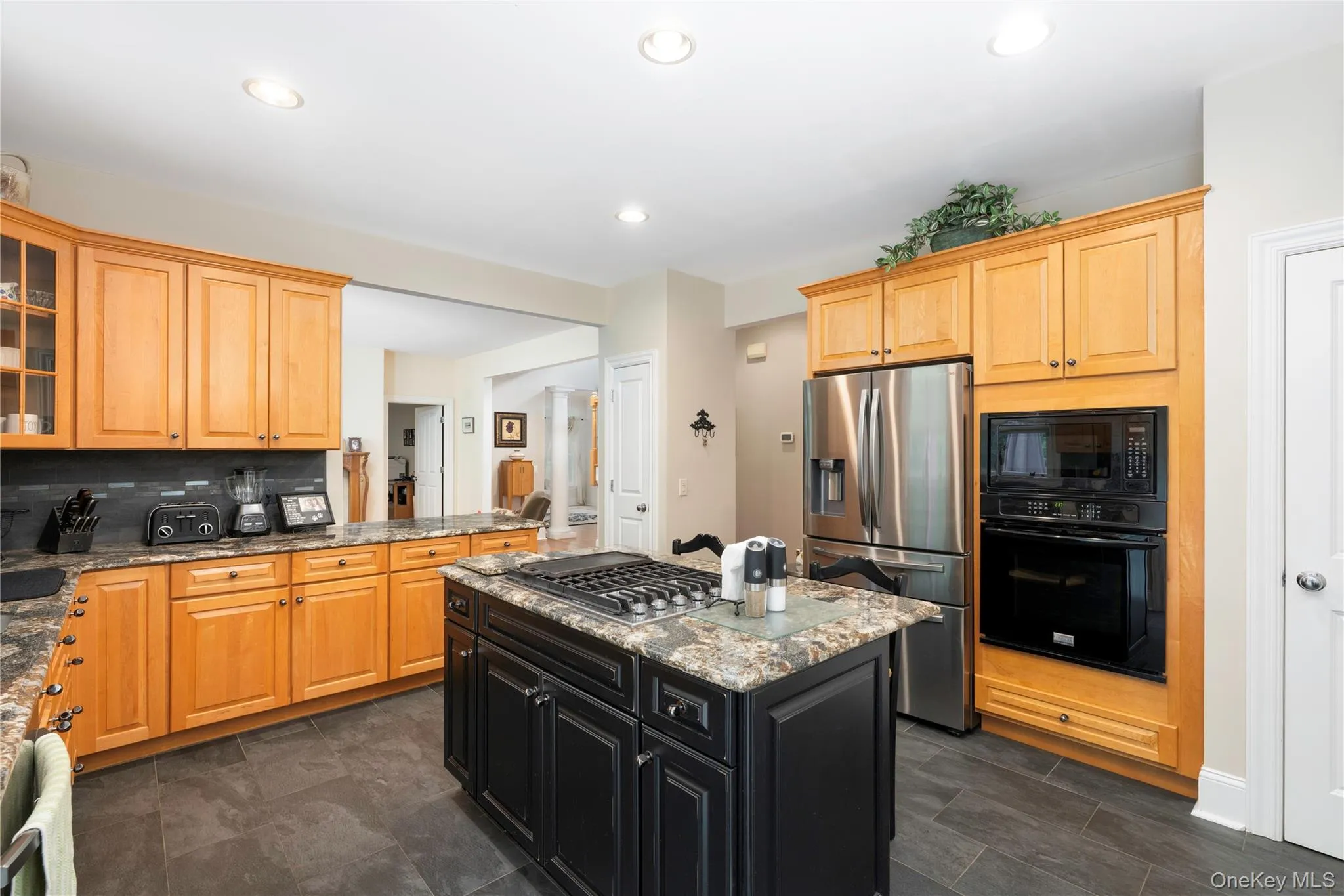 Kitchen featuring a black island with gas cooktop, granite countertops, wood cabinetry, stainless steel refrigerator, and dark tile flooring Kitchen featuring a black island with gas cooktop, granite countertops, wood cabinetry, stainless steel refrigerator, and dark tile flooring