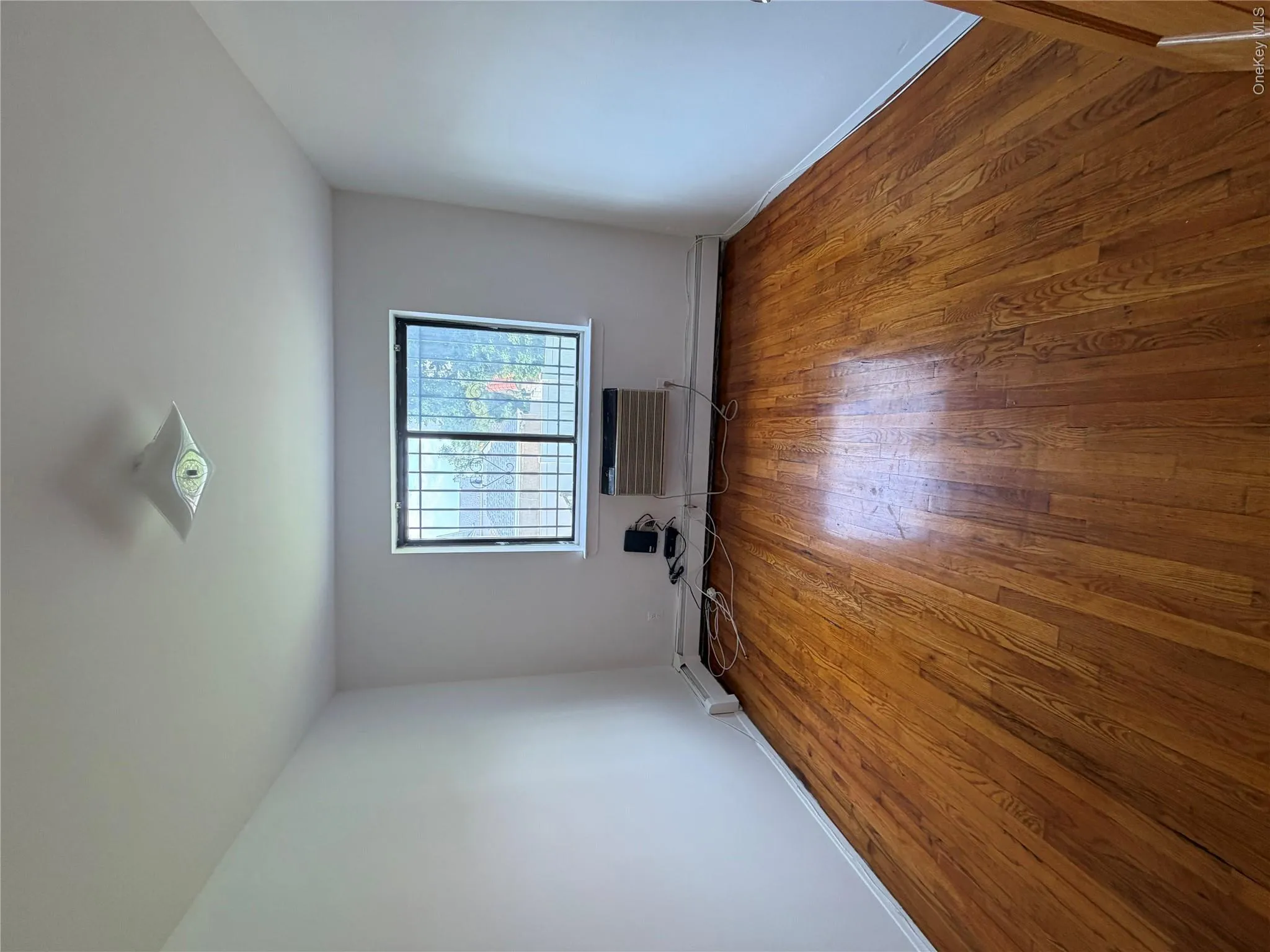 Hardwood flooring throughout, featuring a window with security bars, a ceiling-mounted light fixture, and white painted walls with baseboard trim A/c unit Hardwood flooring throughout, featuring a window with security bars, a ceiling-mounted light fixture, and white painted walls with baseboard trim A/c unit