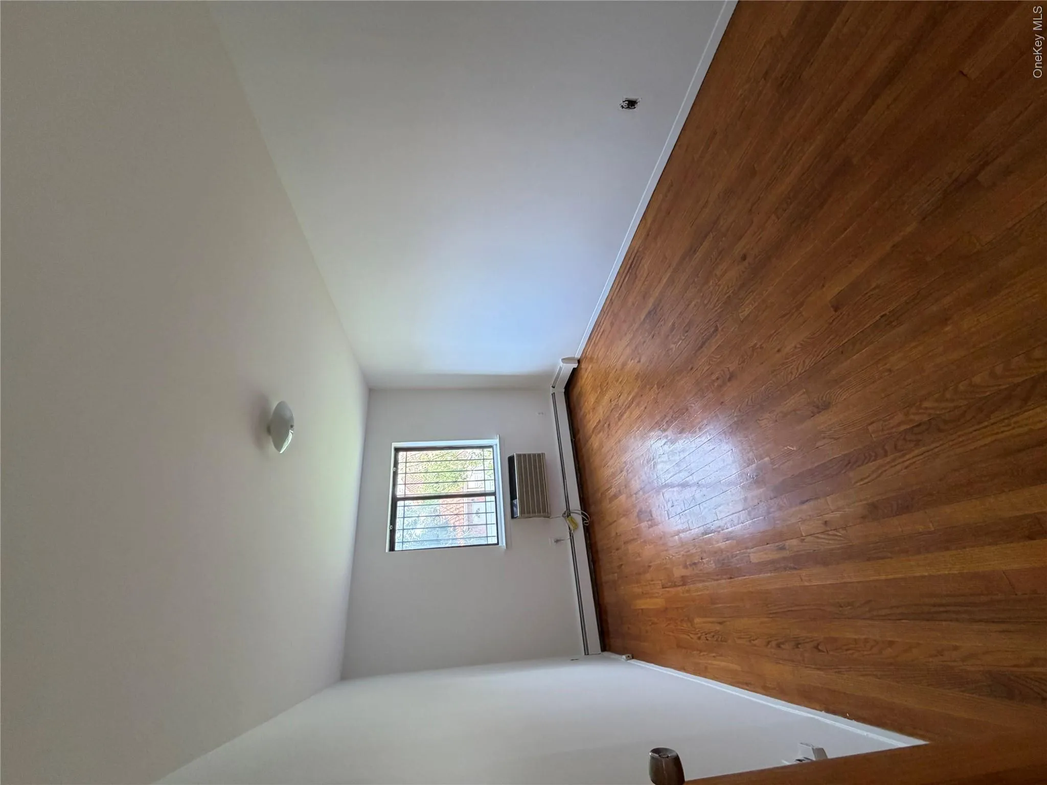 Room featuring wood-finish flooring, white painted walls, and a window with security bars a/c unit Room featuring wood-finish flooring, white painted walls, and a window with security bars a/c unit