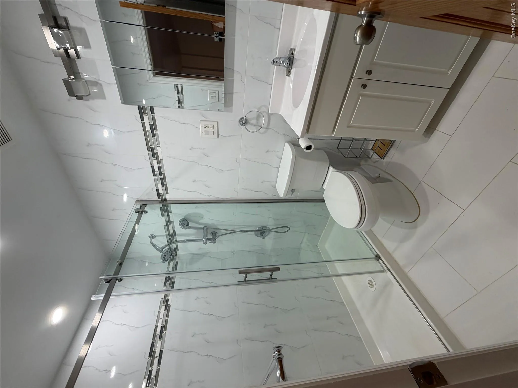 Bathroom featuring a glass-enclosed shower with dual shower heads, white marble-look wall tiling with a decorative accent band, a white vanity with a built-in sink, and a mirrored medicine cabinet Bathroom featuring a glass-enclosed shower with dual shower heads, white marble-look wall tiling with a decorative accent band, a white vanity with a built-in sink, and a mirrored medicine cabinet