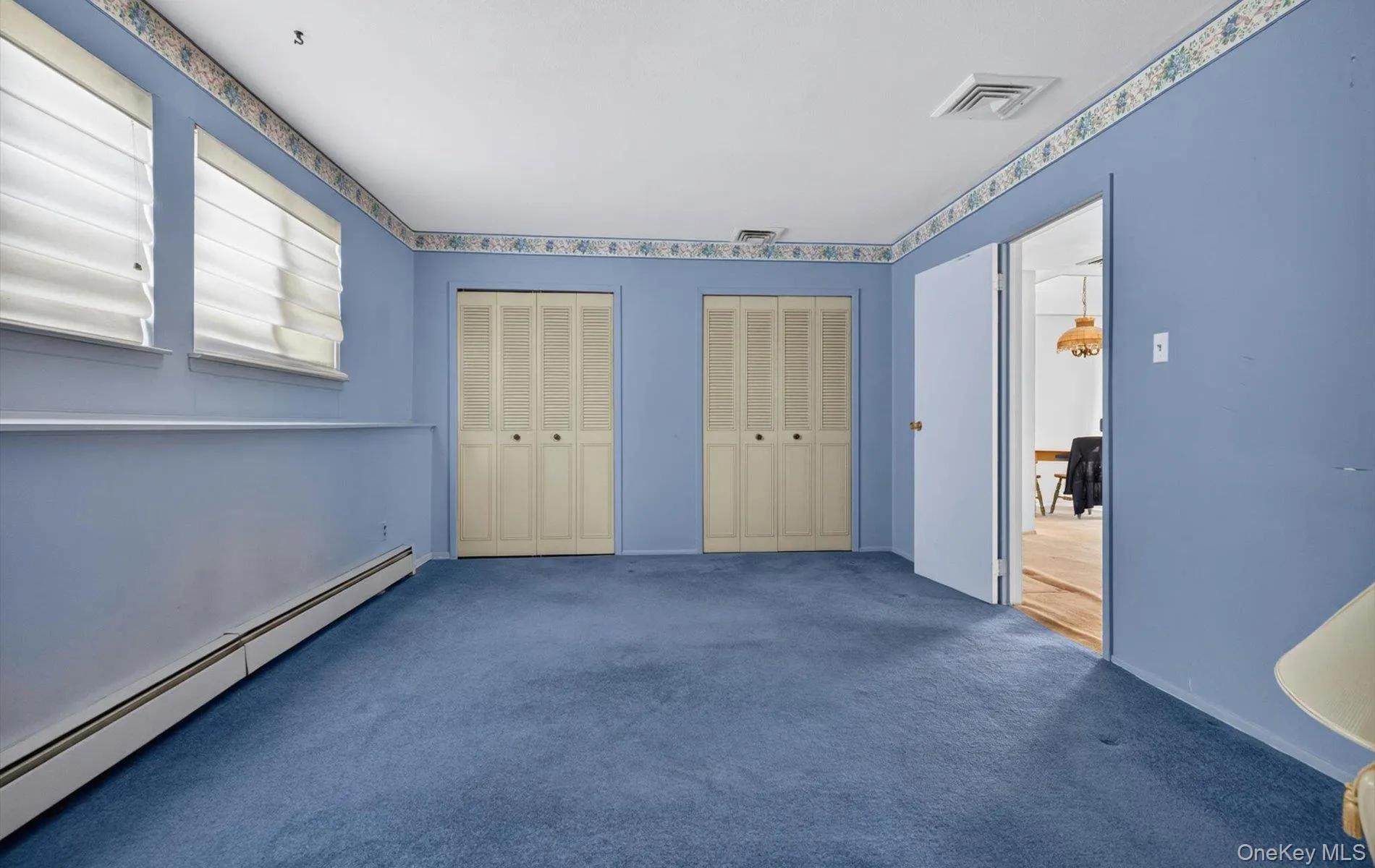 Spacious room featuring blue carpeting, two sets of louvered bi-fold closet doors, and two windows with blinds Spacious room featuring blue carpeting, two sets of louvered bi-fold closet doors, and two windows with blinds