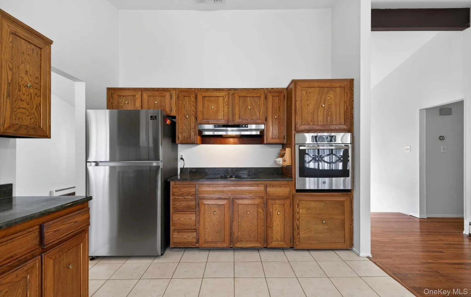 Kitchen featuring wood cabinetry, stainless steel refrigerator, built-in oven, dark countertops, and tile flooring Kitchen featuring wood cabinetry, stainless steel refrigerator, built-in oven, dark countertops, and tile flooring