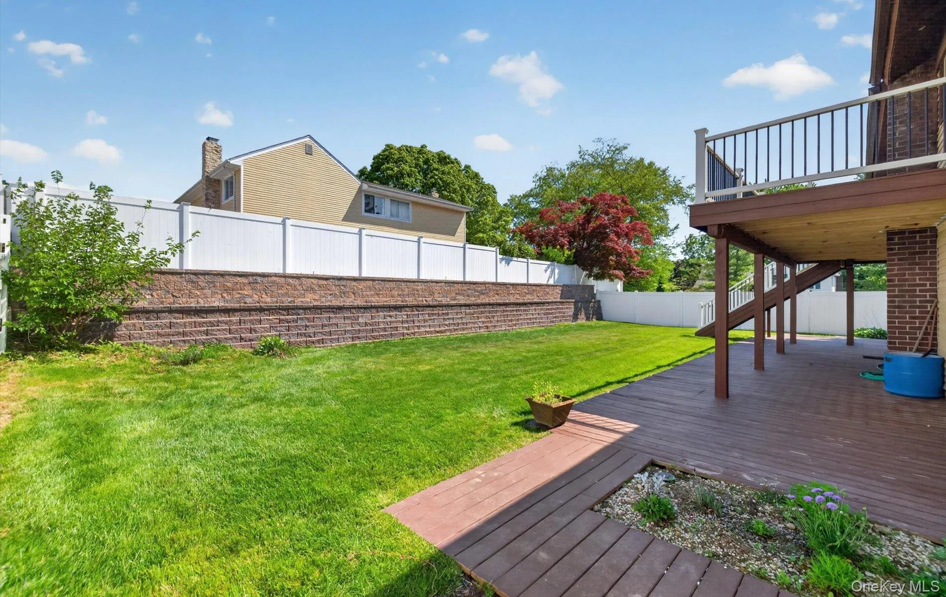 Expansive backyard featuring a large elevated wood-finish deck with railing, a ground-level wood-finish patio area, and a green lawn bordered by a block retaining wall and white privacy fencing Expansive backyard featuring a large elevated wood-finish deck with railing, a ground-level wood-finish patio area, and a green lawn bordered by a block retaining wall and white privacy fencing