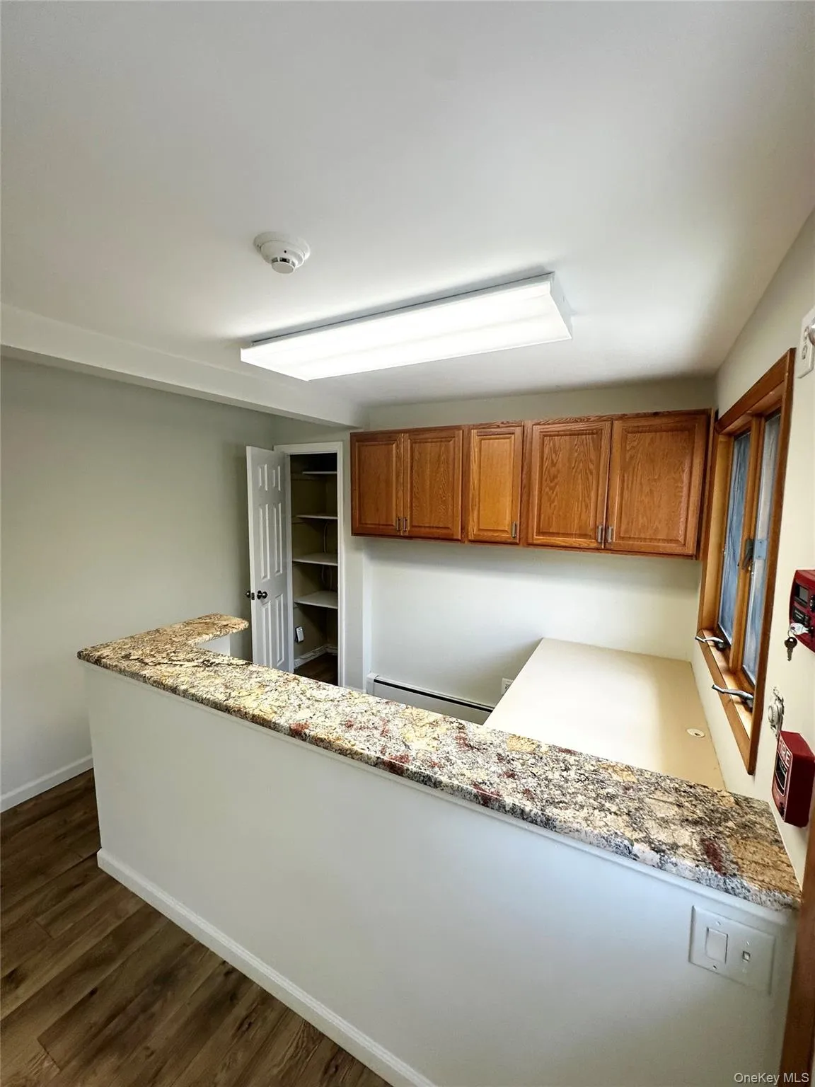 Kitchen with brown cabinetry, dark wood-type flooring, a baseboard radiator, built in desk, and light stone countertops Kitchen with brown cabinetry, dark wood-type flooring, a baseboard radiator, built in desk, and light stone countertops
