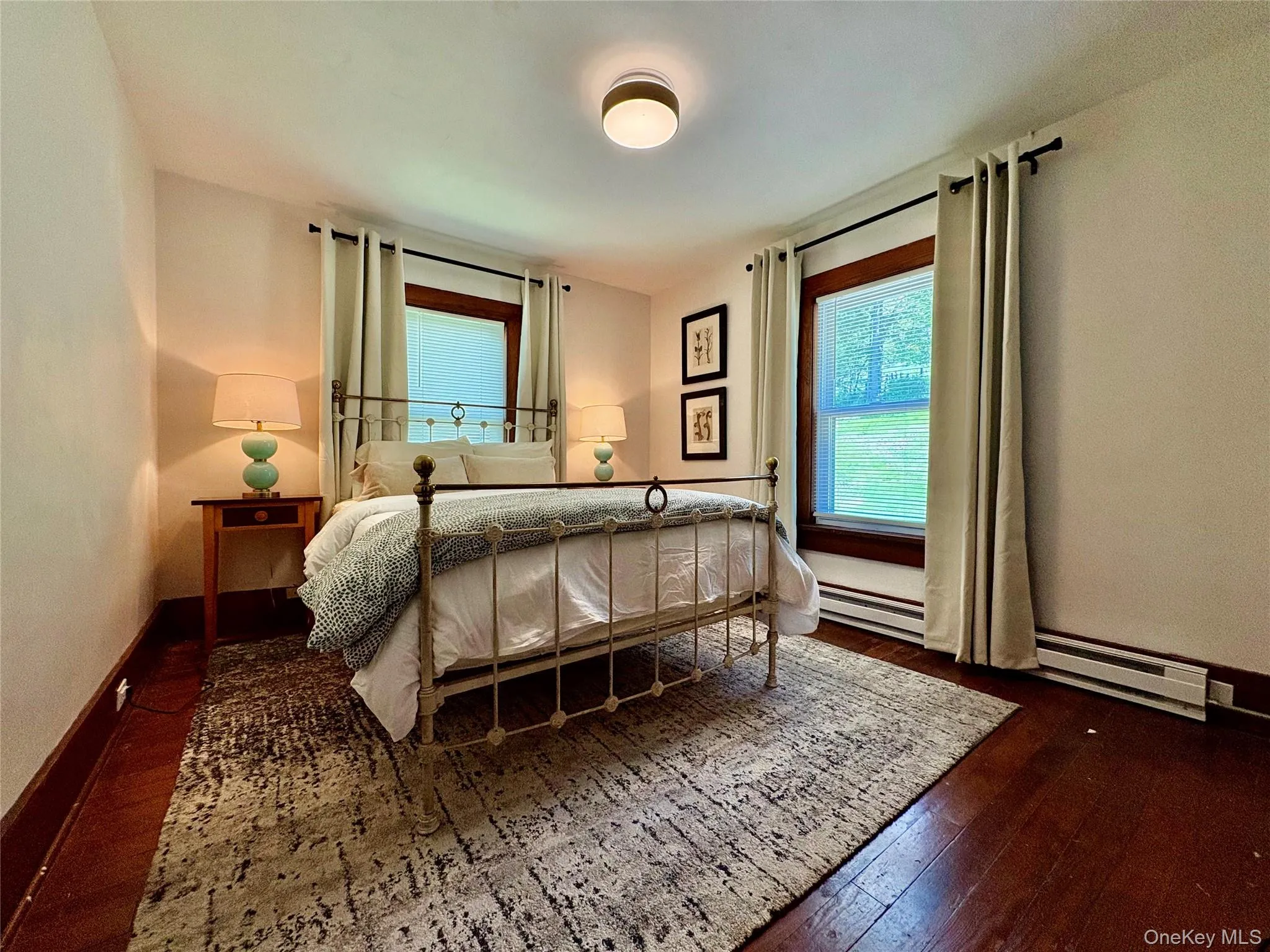 Bedroom featuring wood-finish flooring, two windows with trim, baseboard heating, and a flush-mount ceiling light Bedroom featuring wood-finish flooring, two windows with trim, baseboard heating, and a flush-mount ceiling light