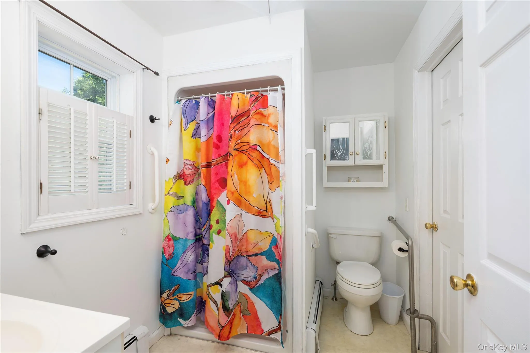 White bathroom featuring a shower with grab bars, toilet with adjacent grab bars, and a window with white louvered shutters White bathroom featuring a shower with grab bars, toilet with adjacent grab bars, and a window with white louvered shutters